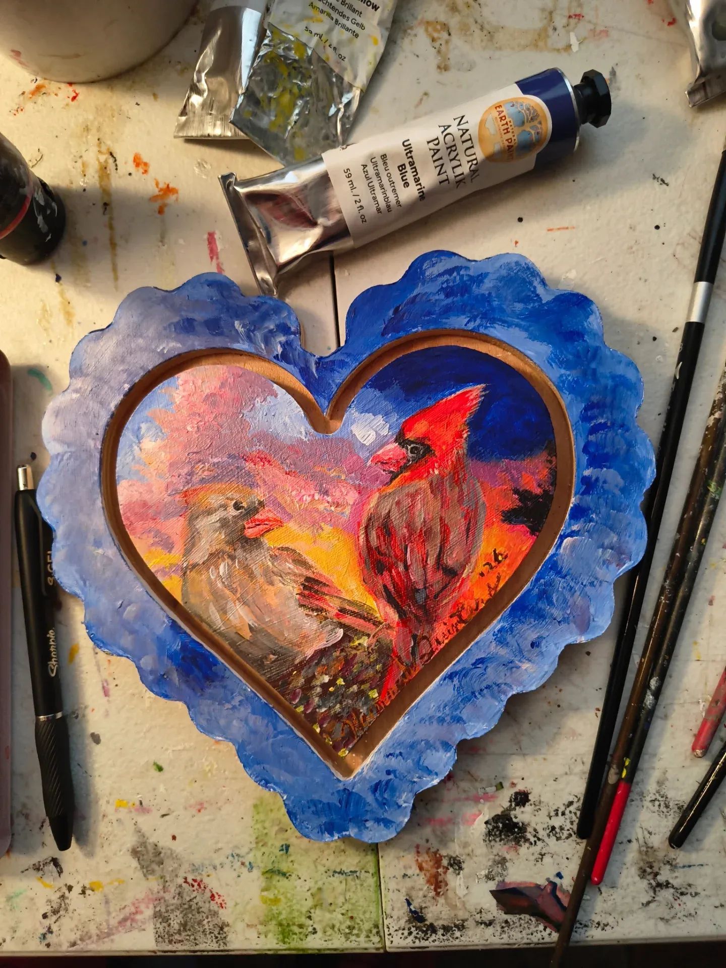 Heart-shaped painting of a red bird on a branch with a pink and orange sky, surrounded by a blue border.