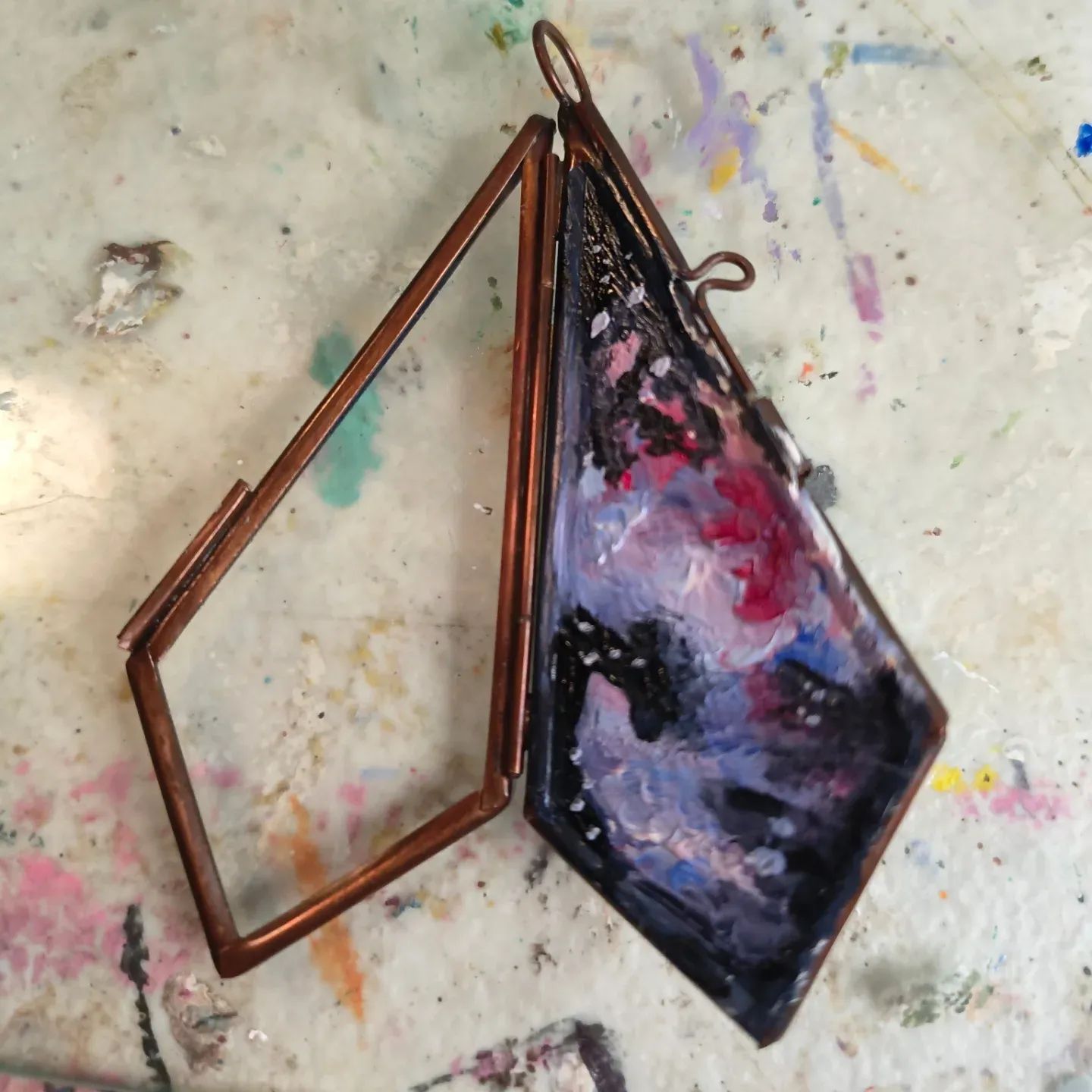 A small, rectangular, painted object with a metal frame, lying on a paint-splattered surface.