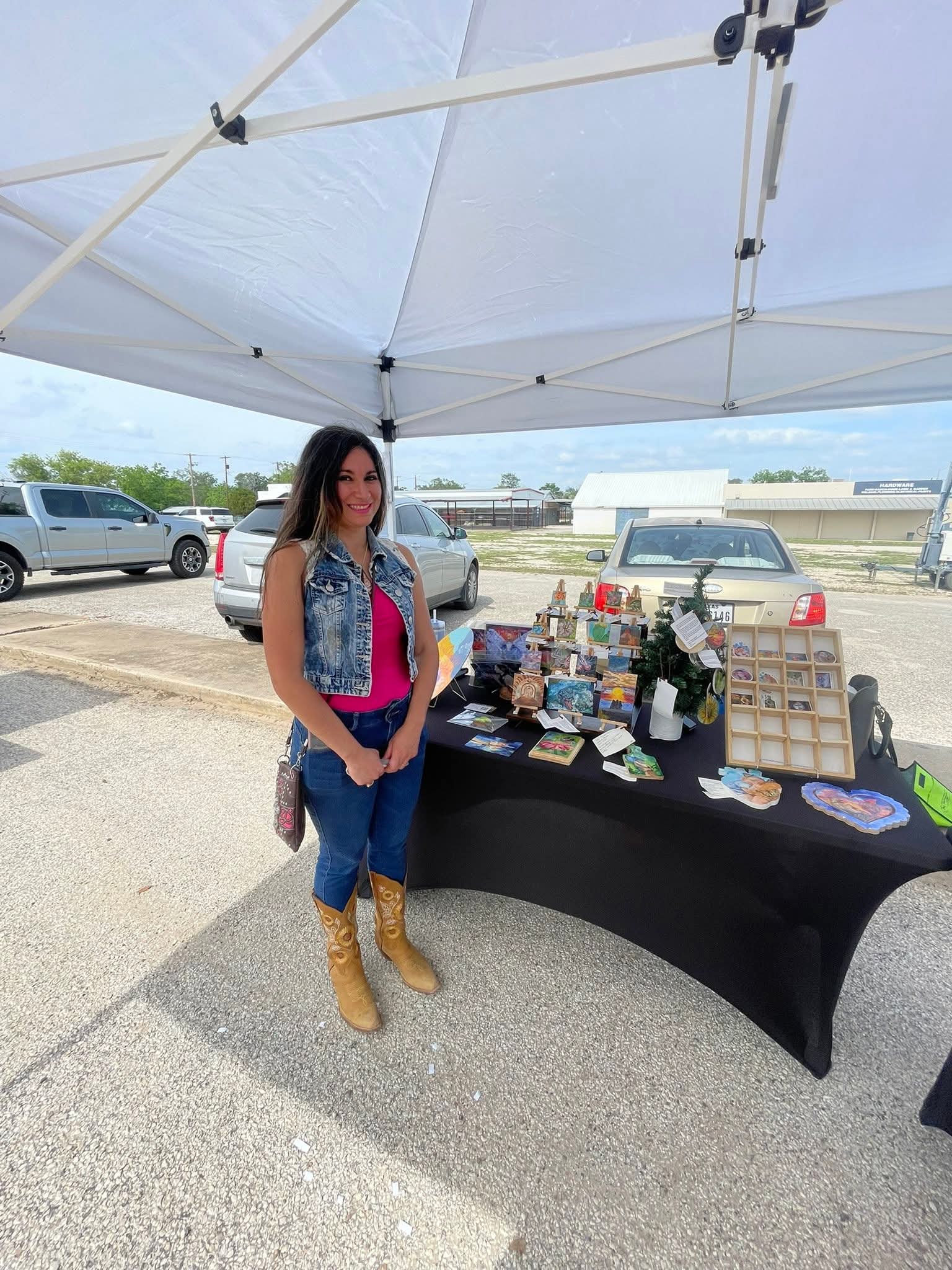 Woman standing next to a table with various items under a white canopy outdoors, with parked cars in the background.