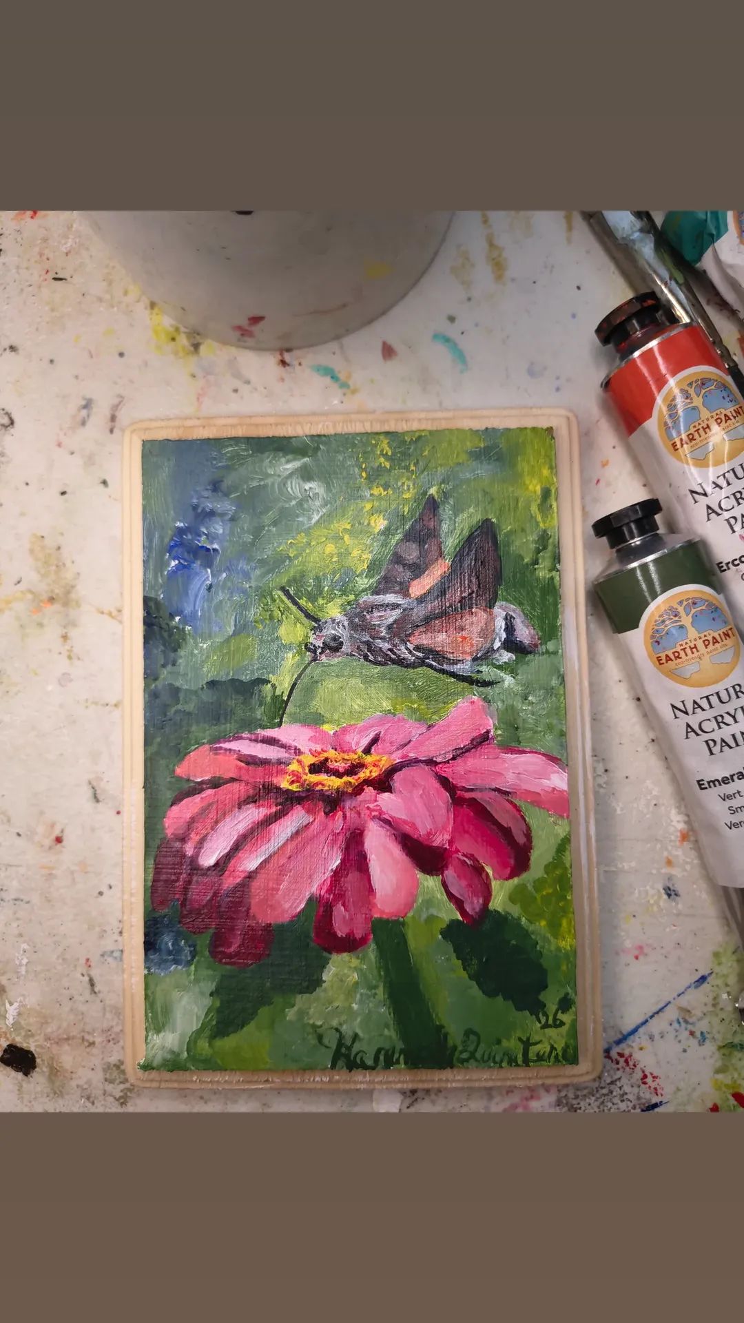 Painting of a pink flower with a butterfly on it, background of green foliage, on an artist's workspace with paint tubes.