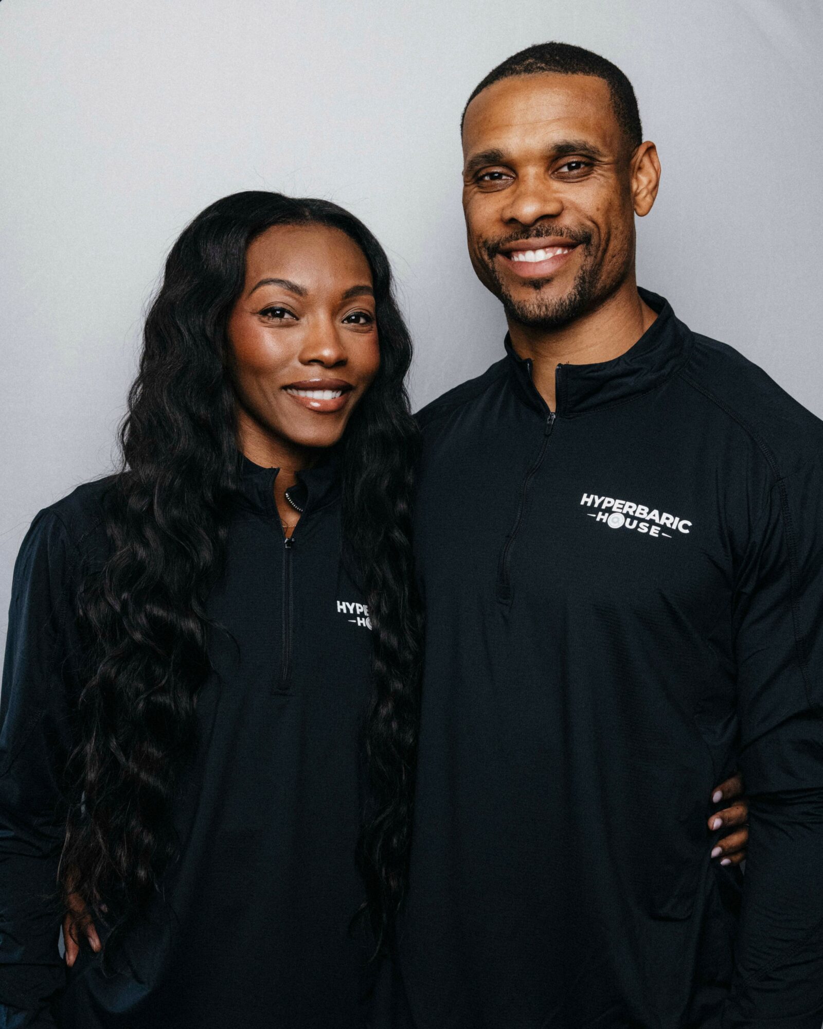 Two people standing close together, smiling, wearing black Hyperbaric House jackets, against a plain light background.