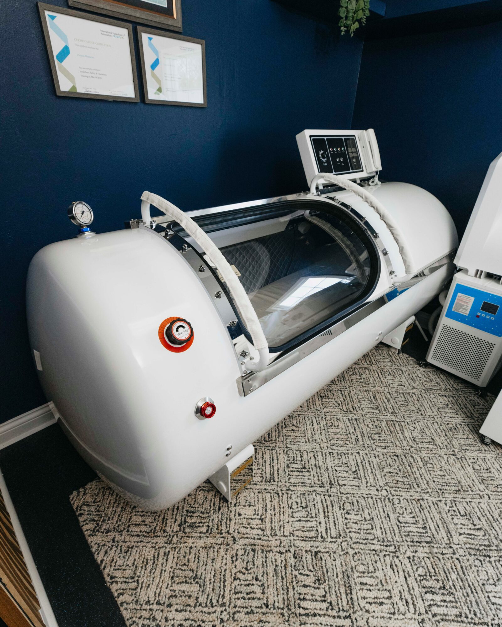 Medical imaging machine with control panel, situated in a room with dark blue walls and framed certificates, on a patterned carpet.