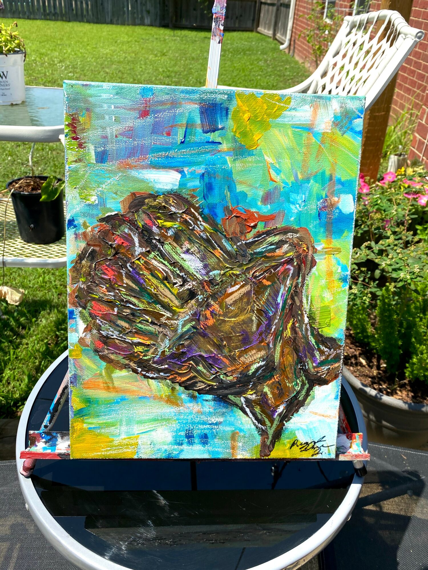 Colorful painting of a hand with vibrant brushstrokes, set outdoors on a stand with garden plants in background.