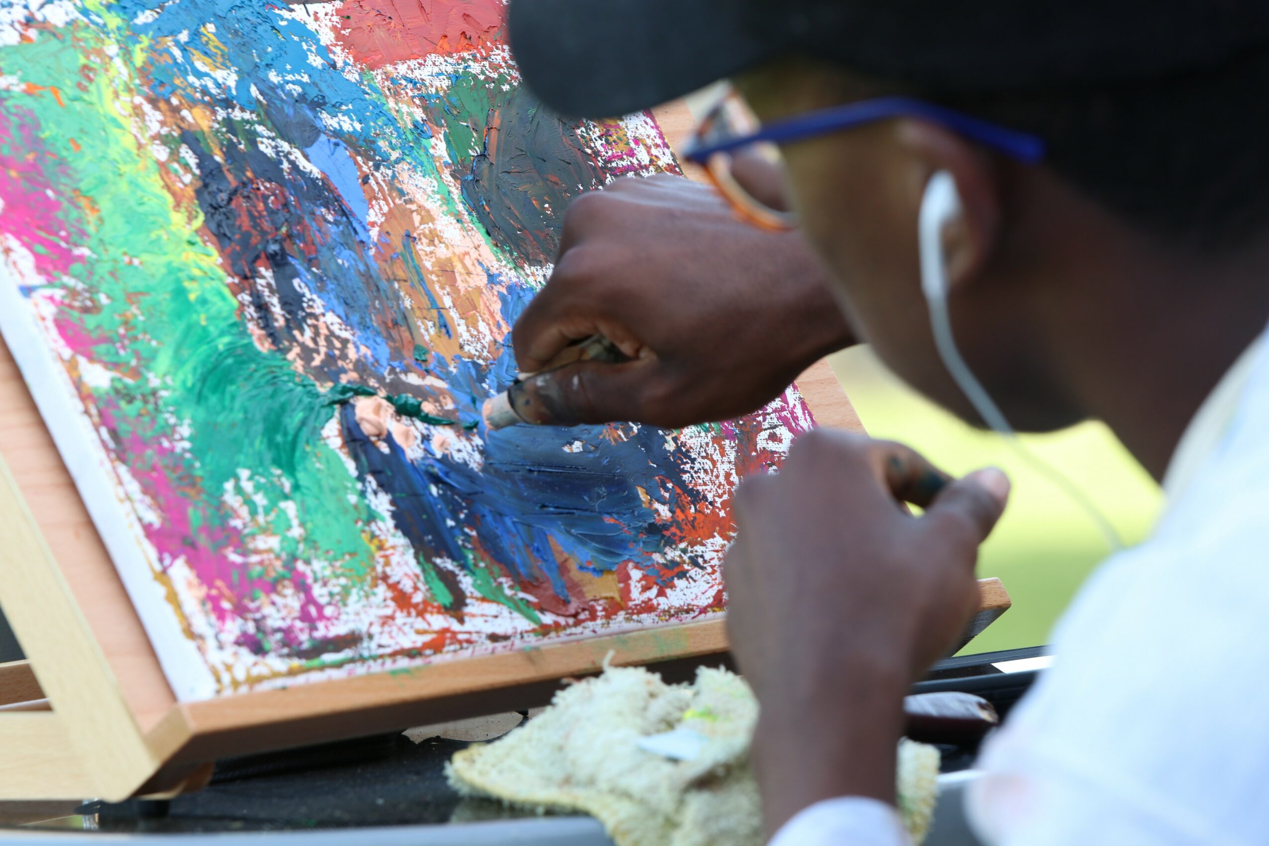 Person painting an abstract colorful artwork with a palette knife, wearing glasses and earphones, close-up view.