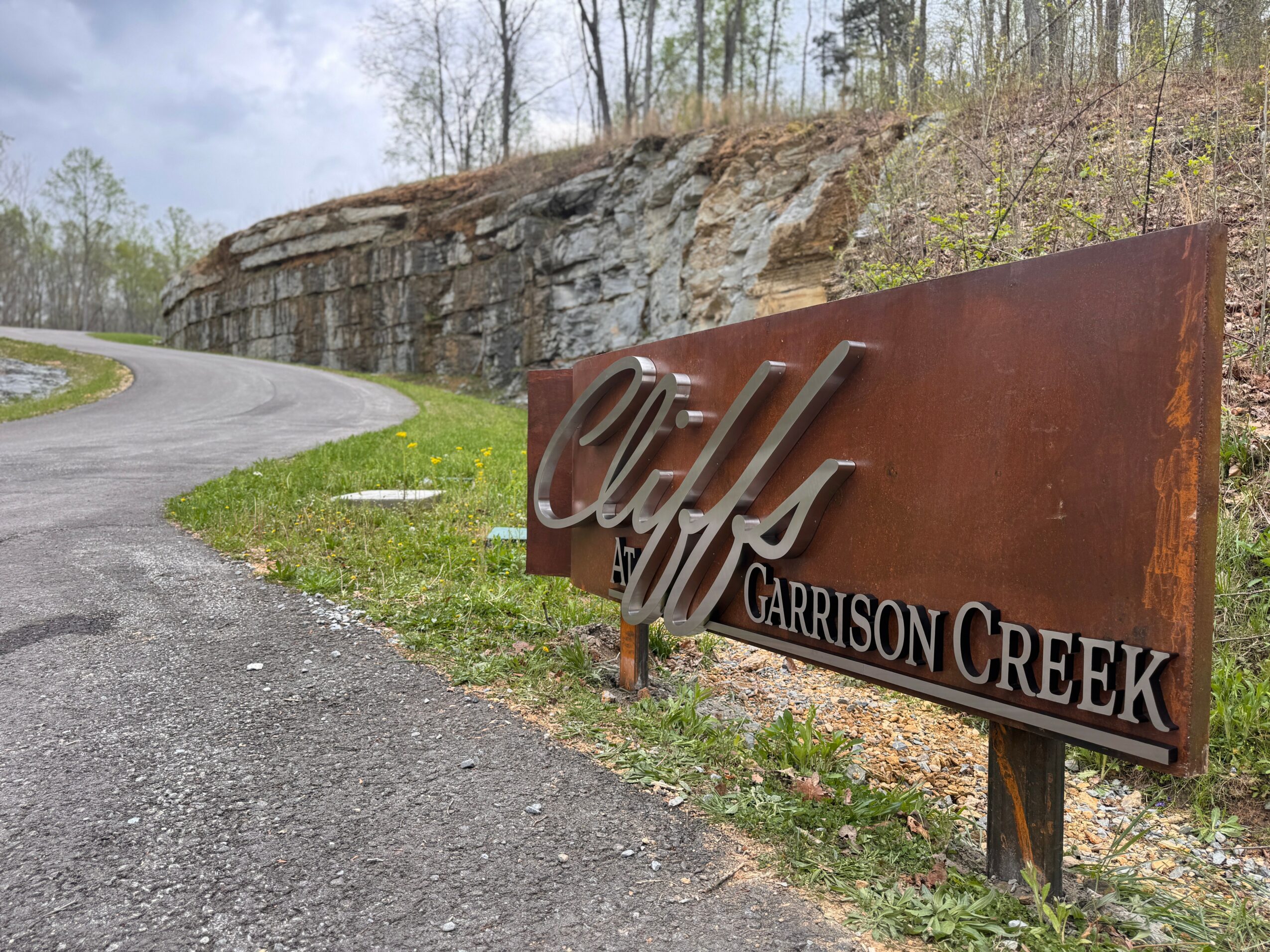 Rusty sign reads 'Chilf at Garrison Creek' beside a curved road with a rocky hillside and trees in the background.