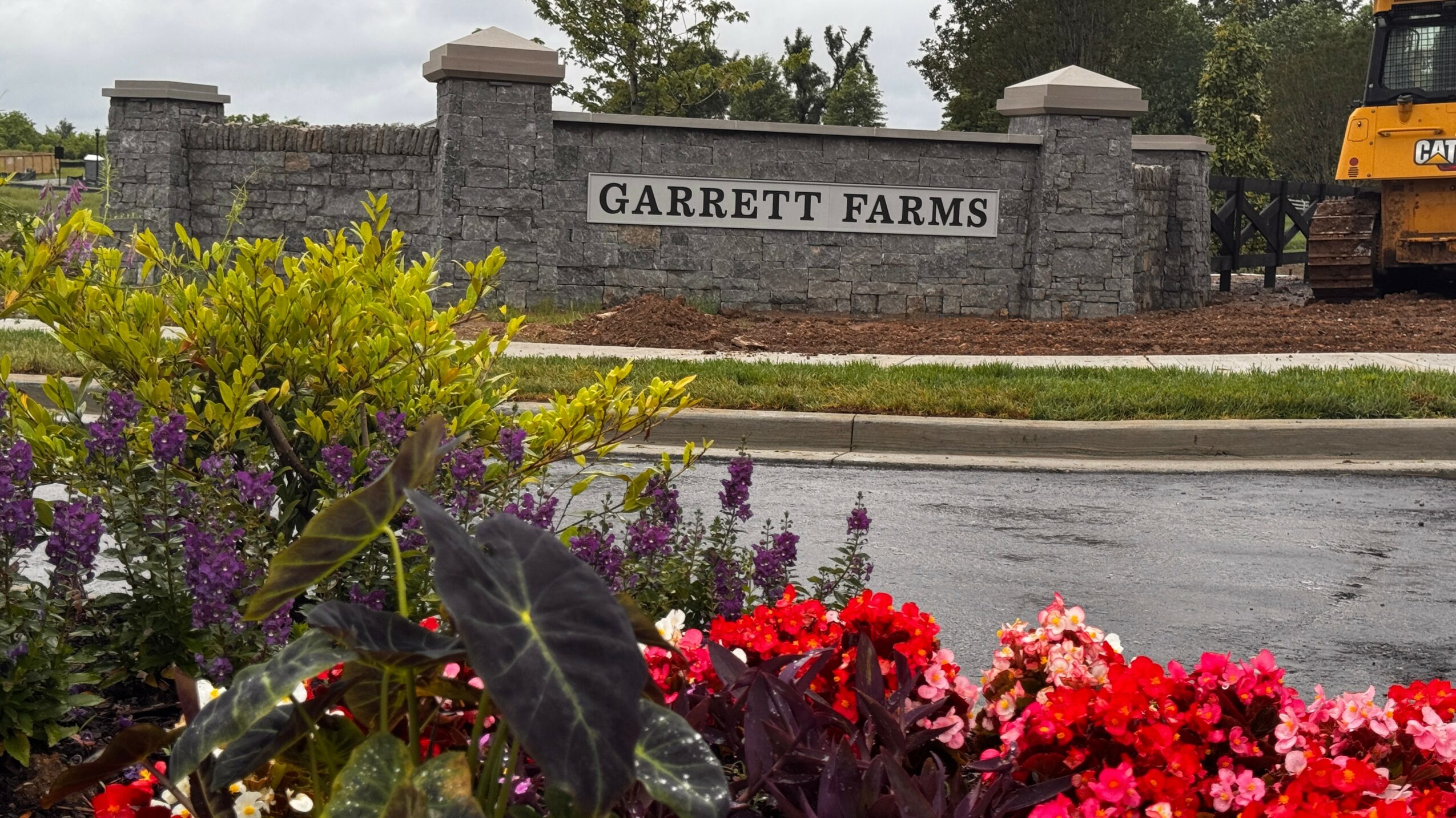 Stone entrance sign reads Garrett Farms, with colorful flowers in foreground and a street in the background.