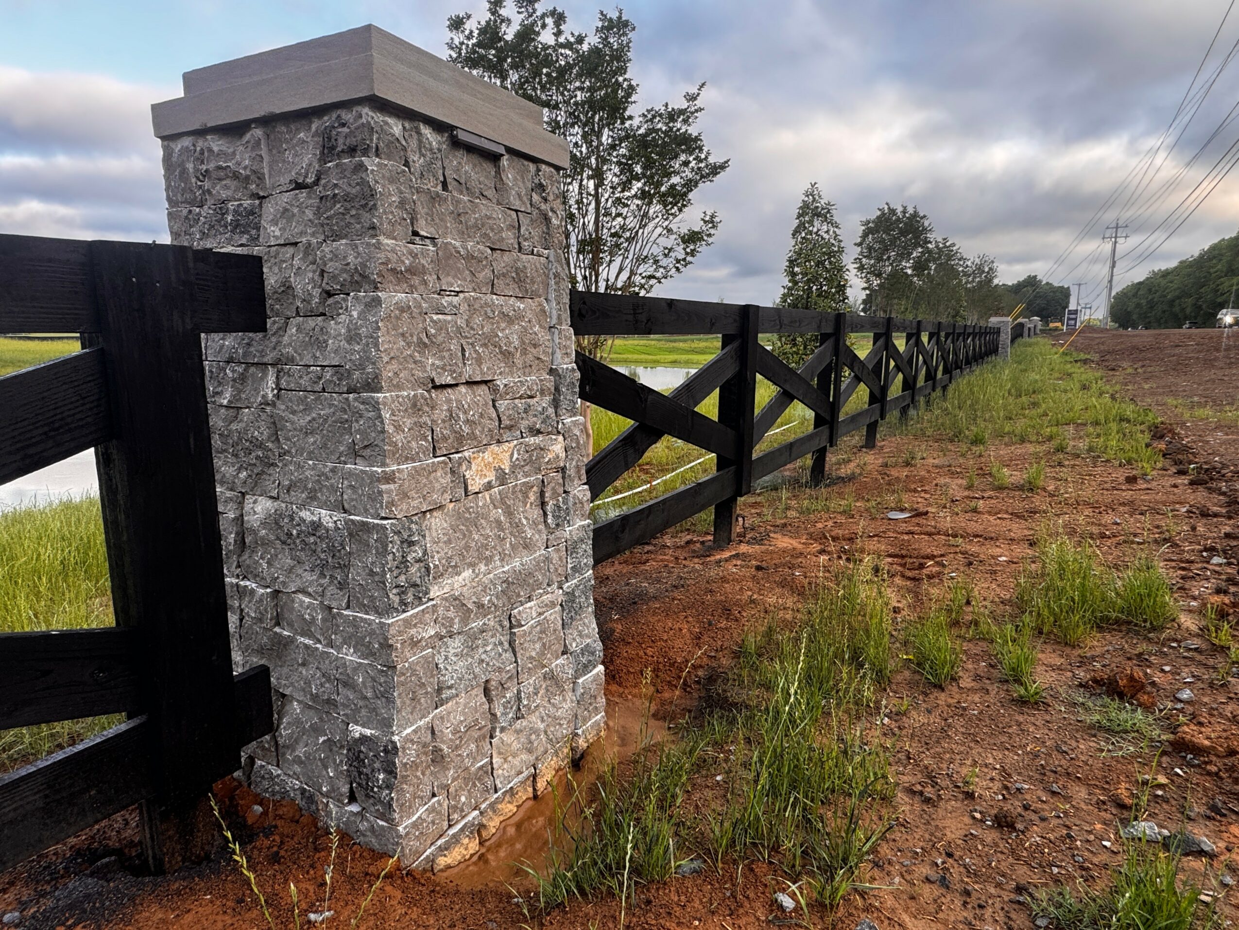 Stone pillar with black wooden fence extending into the distance, grassy and dirt area, cloudy sky.