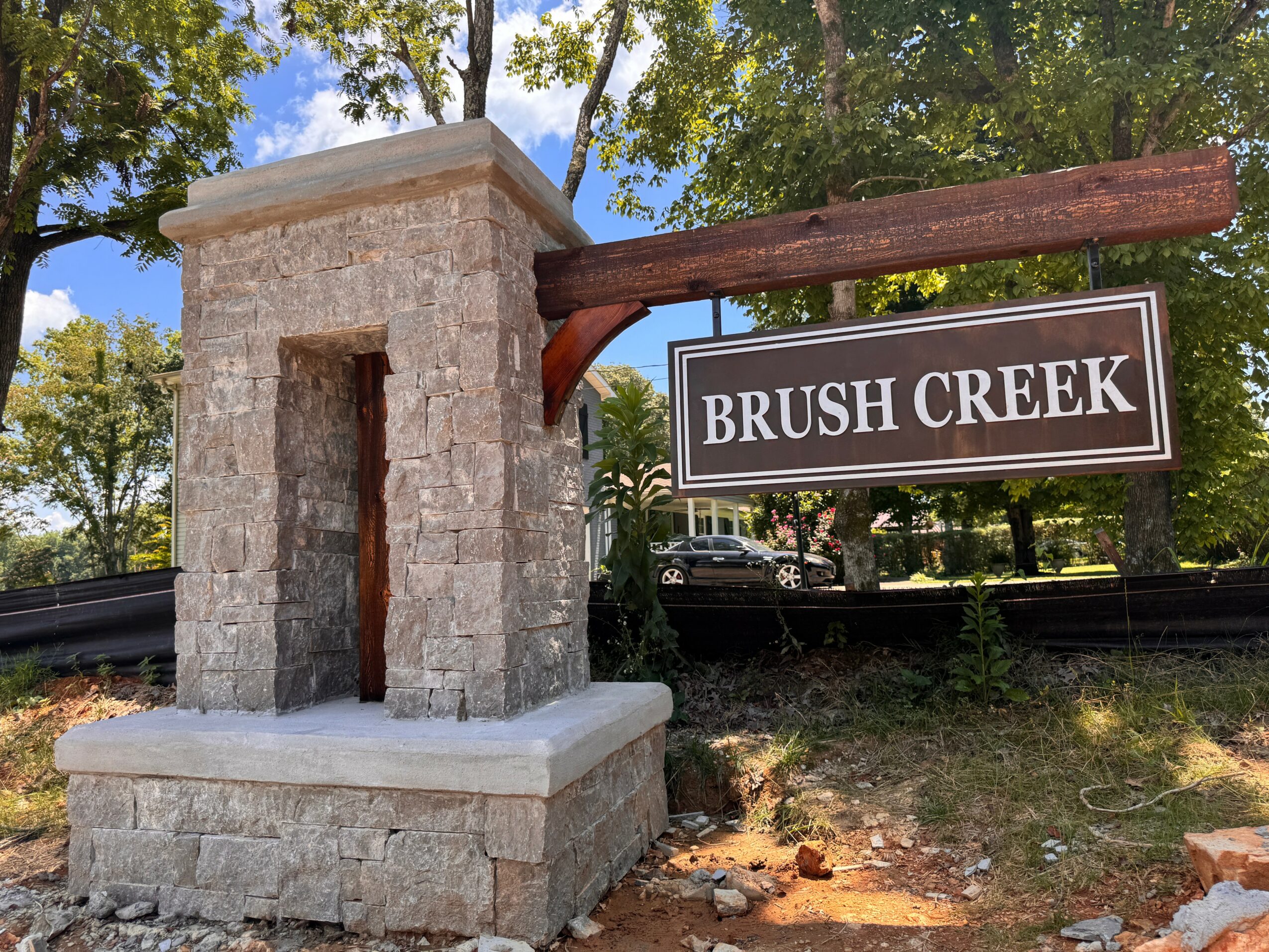 Stone structure with a sign reading 'Brush Creek' hanging from a wooden beam, trees and sky in background.