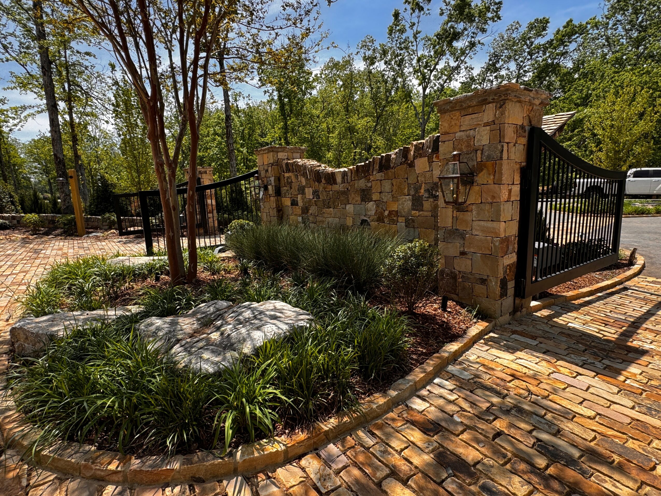Stone wall with black gate, brick pathway, trees, and landscaped plants in a garden setting.