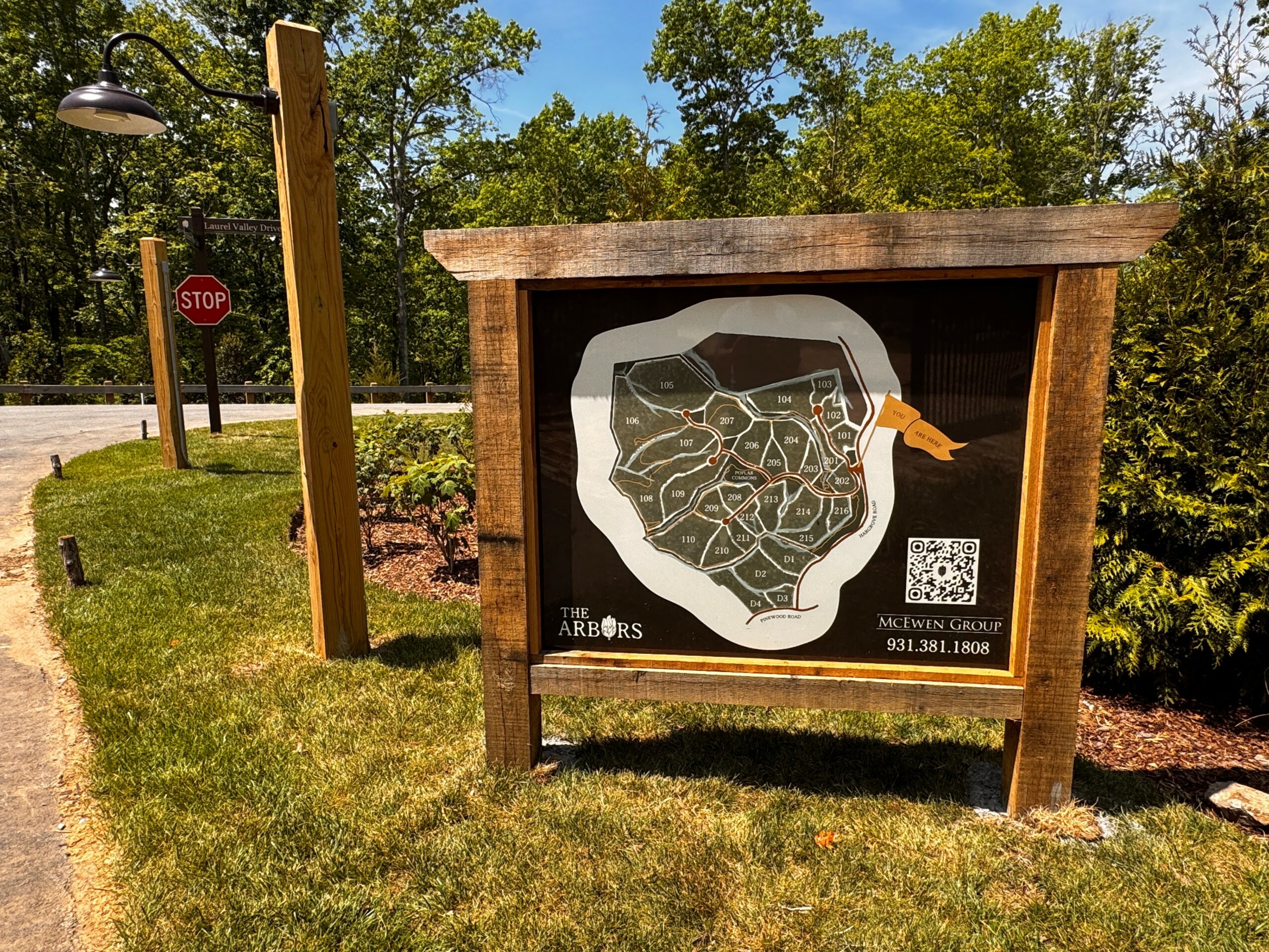 Outdoor informational sign with a map, surrounded by trees and grass, near a stop sign and street lamp.