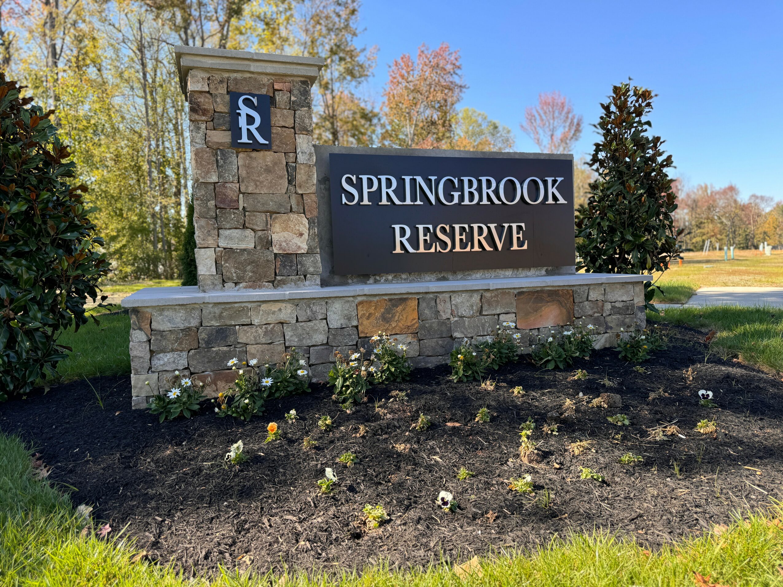Stone sign with blue and white logo, reading 'Springbrook Reserve,' surrounded by a flower bed and trees in background.