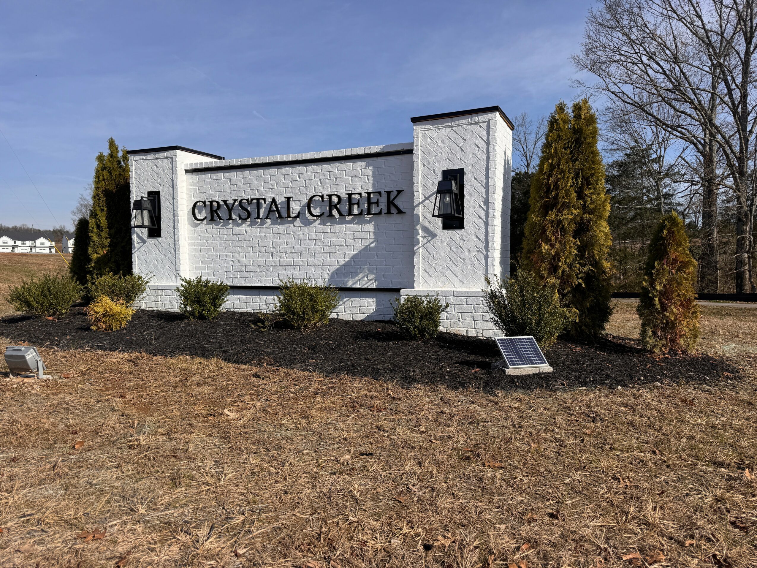 A white sign with black text reads 'CRYSTAL CREEK' and is surrounded by bushes and trees under a blue sky.