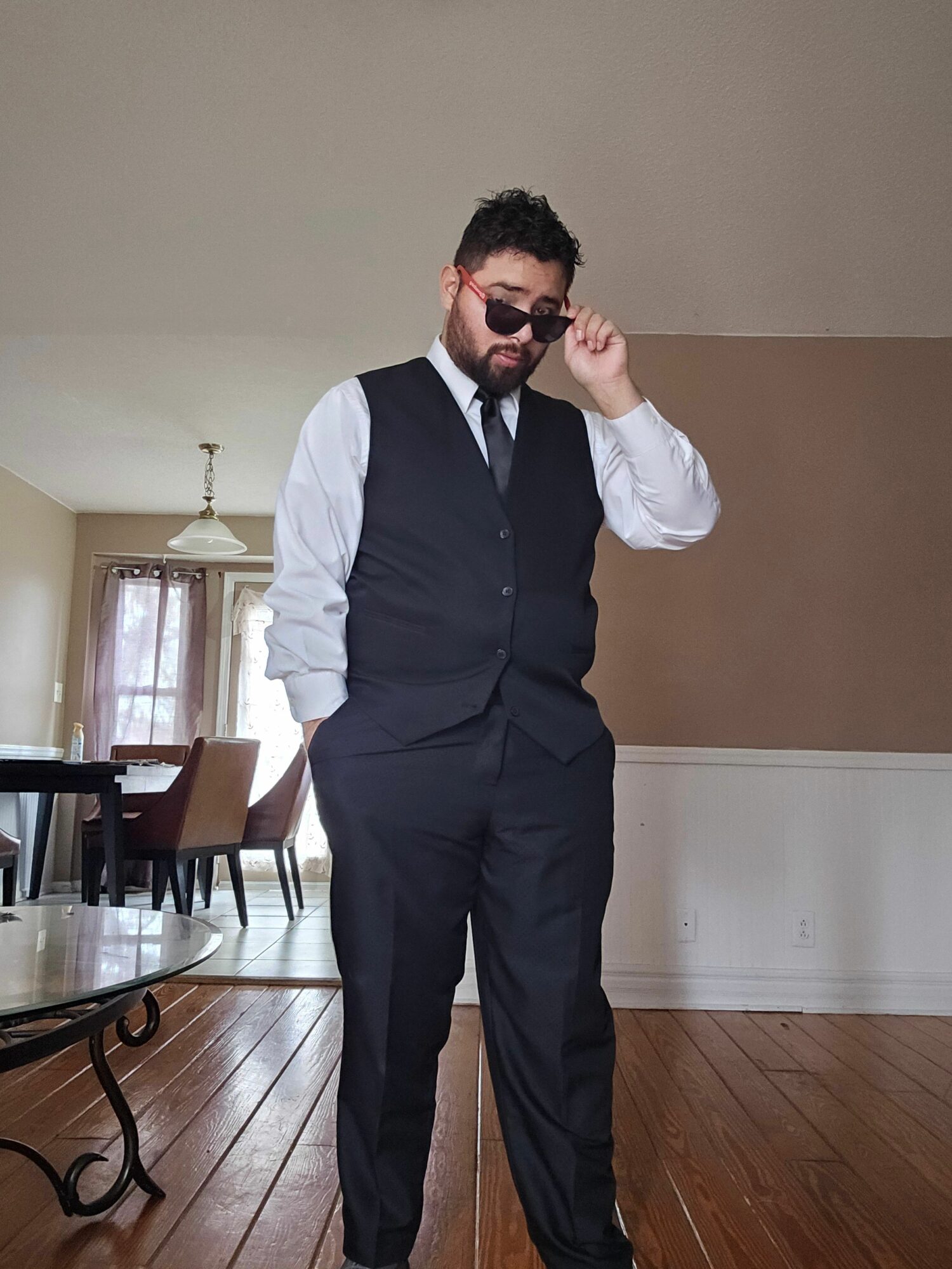 Man in a suit and sunglasses standing in a room with wooden floor and a table, holding a phone to his ear.