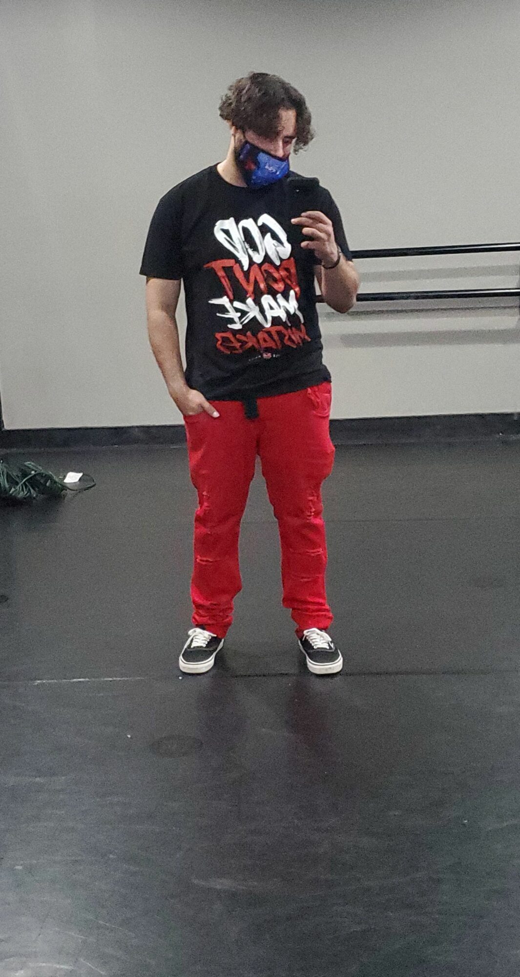 Person standing in a room, wearing a black T-shirt, red pants, sneakers, and a face mask, taking a mirror selfie.