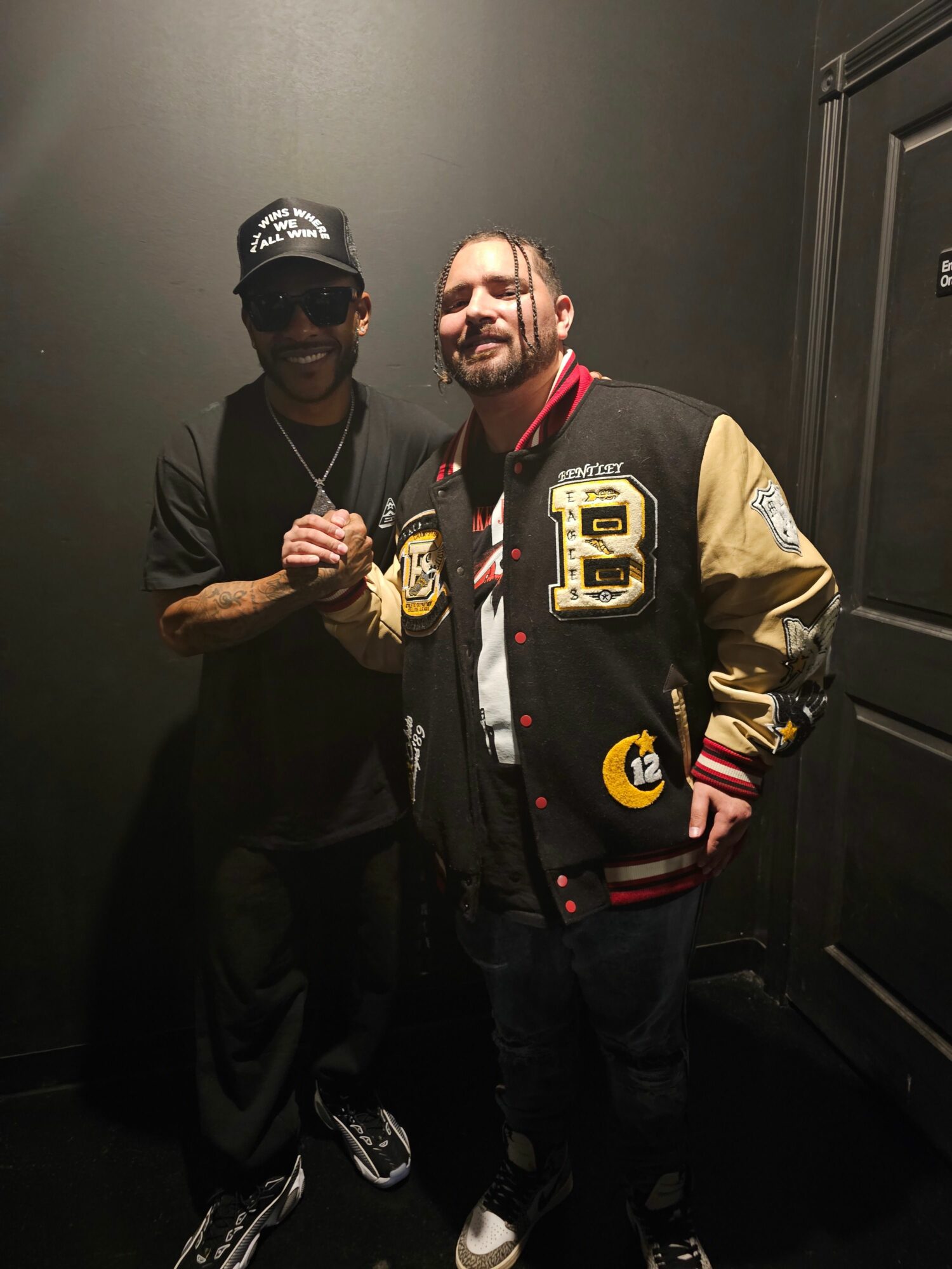 Two people standing indoors, one wearing a varsity jacket and the other in a hoodie and cap, holding a drink.