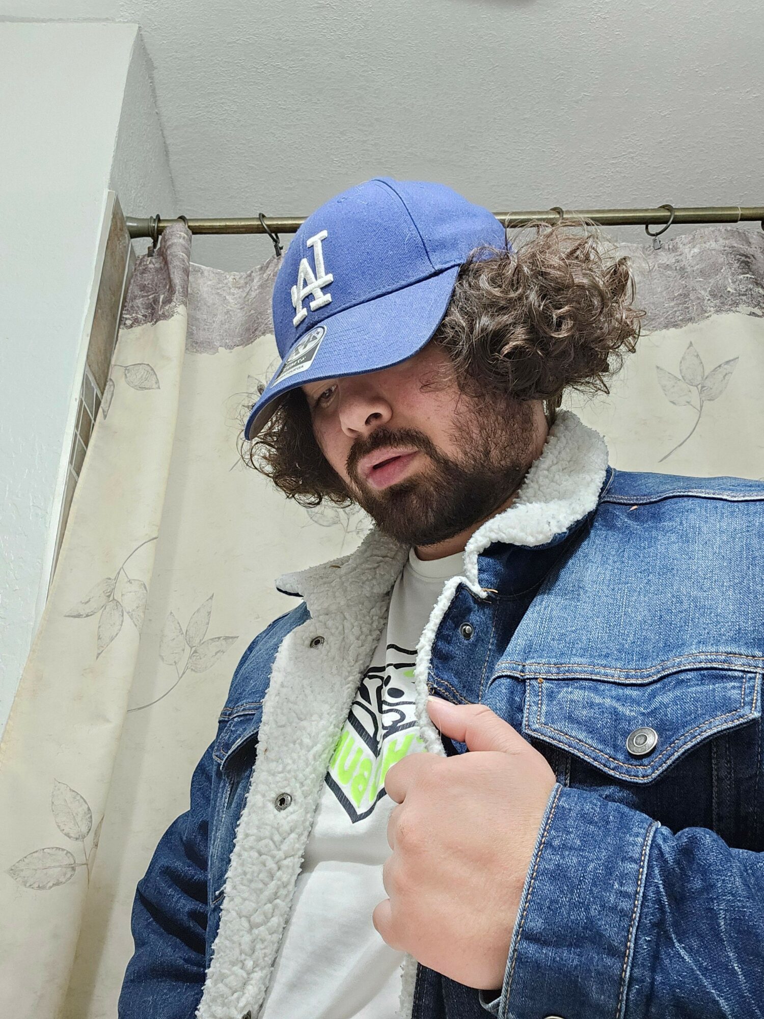 Person with curly hair wearing a blue cap and denim jacket, leaning over a surface, smiling.