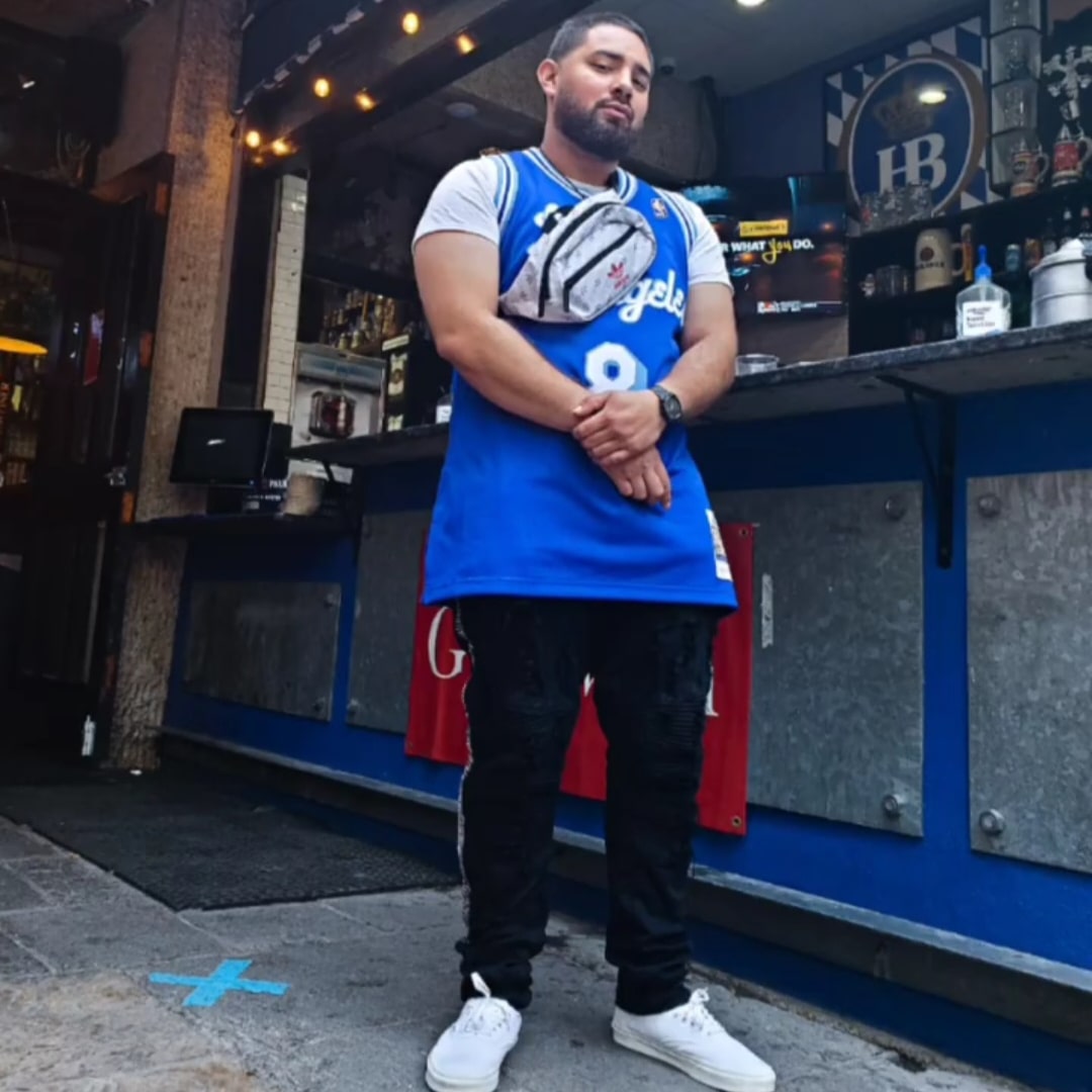 Man with beard in blue sports jersey and black pants stands outside a bar, arms crossed, holding a small bag.