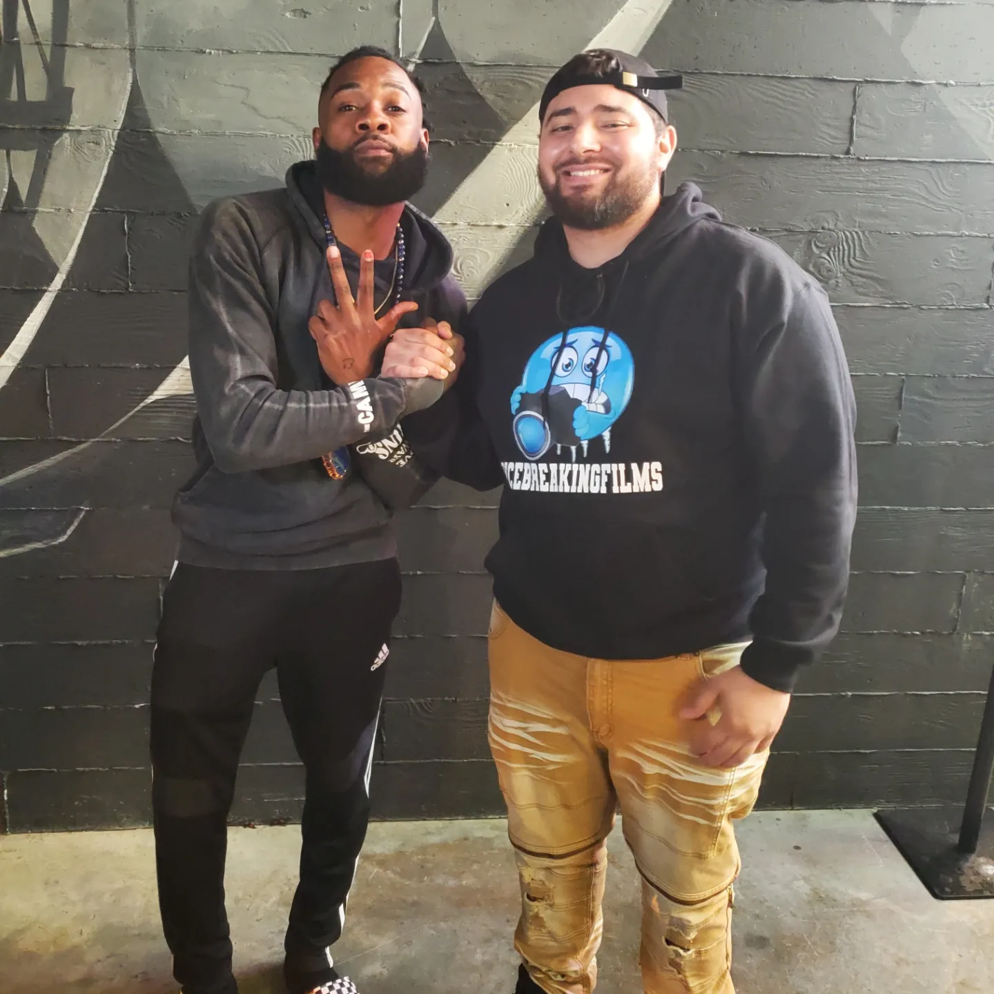 Two men standing against a dark brick wall, smiling, one with a beard and the other wearing a hoodie, casual pose.