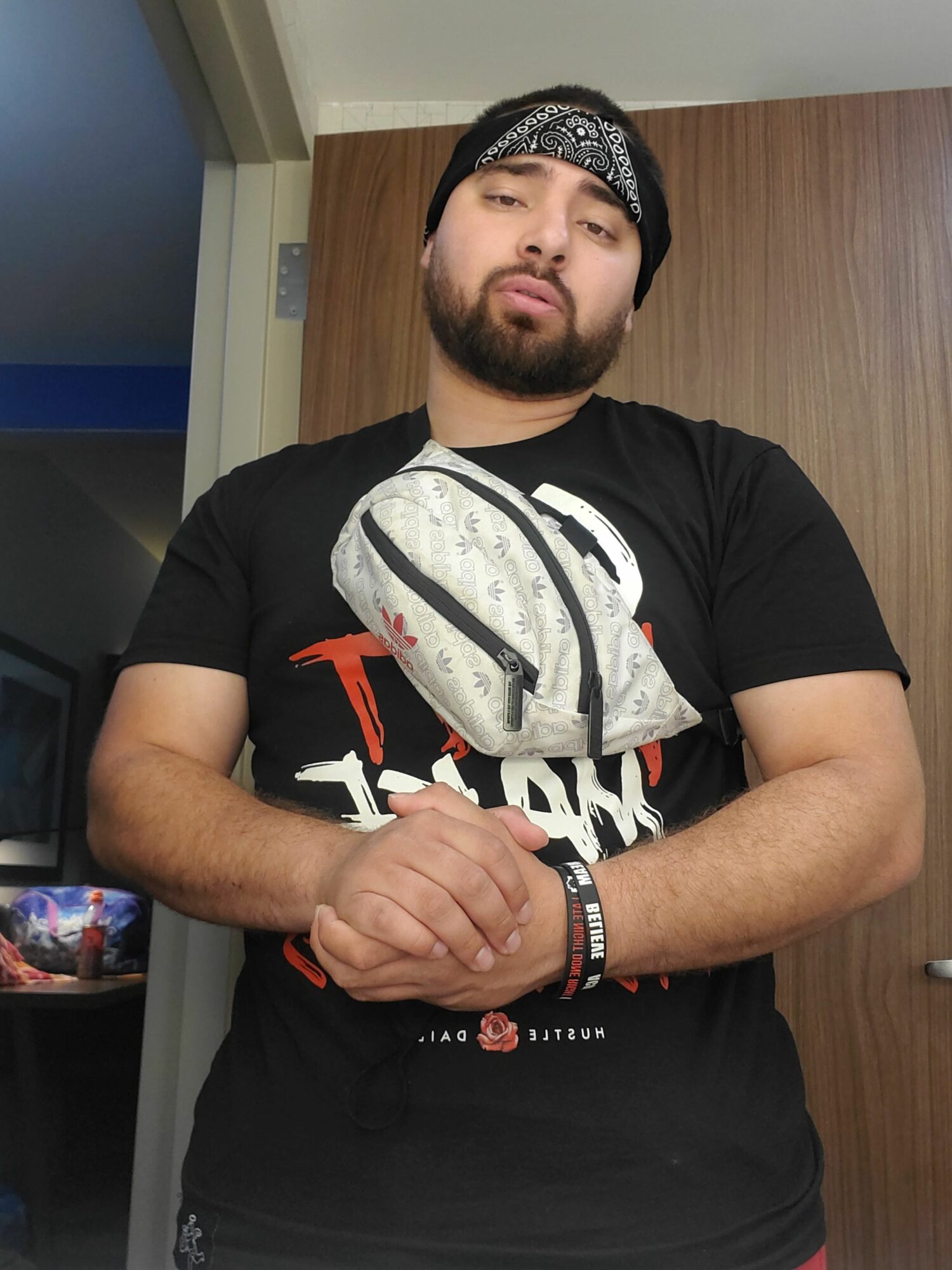 Man with a beard and headband, wearing a black T-shirt, standing indoors with a wooden door behind him.