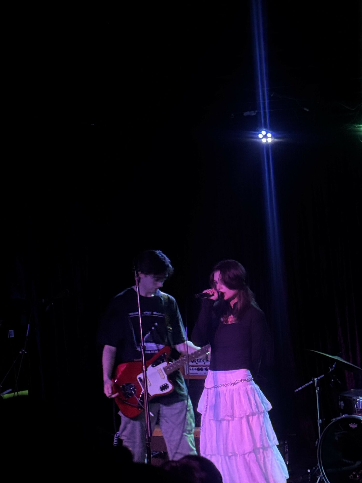 Two people on stage, one playing guitar, the other singing into a microphone, with a dark background and bright light above.
