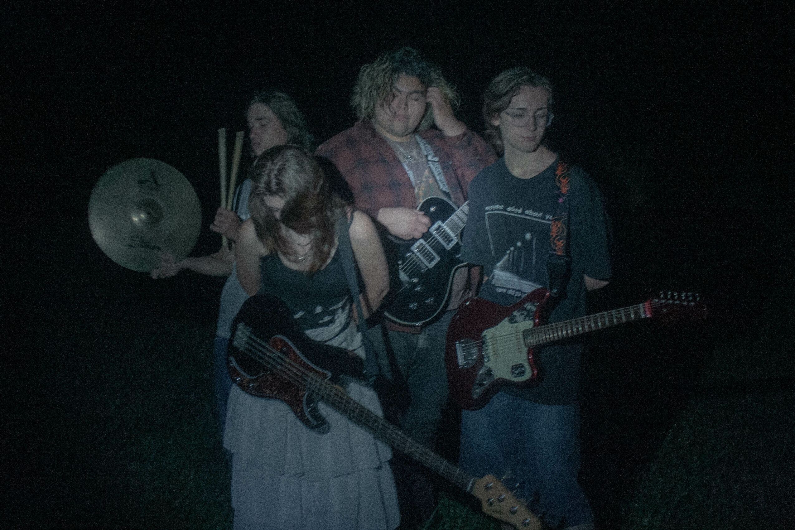 Four people playing guitars and drums in a dark setting, with one person holding a cymbal, all focused on music.