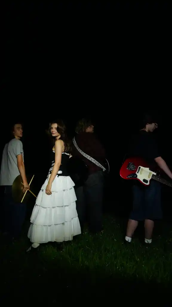 Four people standing outdoors at night, one woman in a white dress, others with musical instruments, dark background