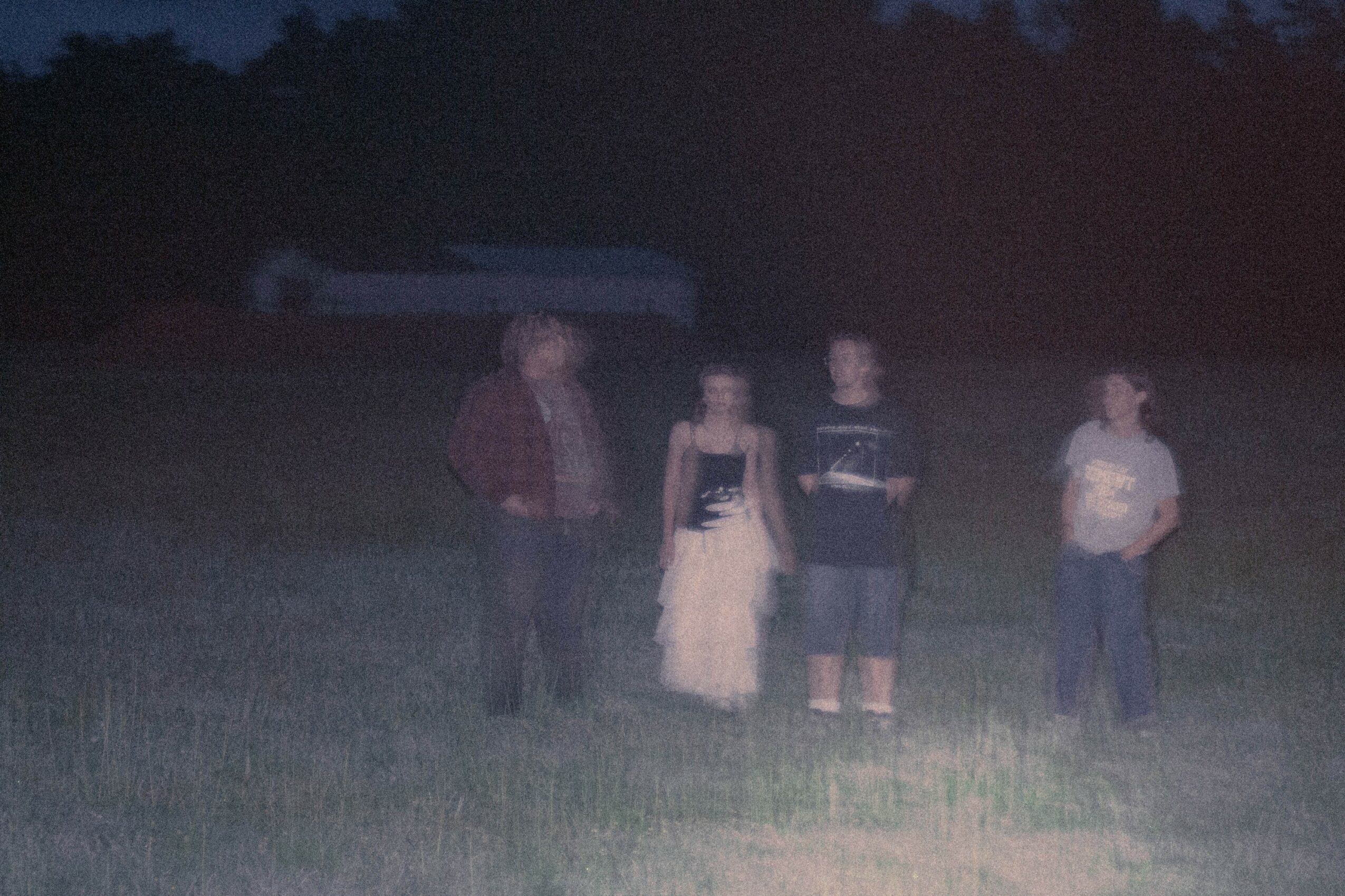 Four people standing on grass in a field at night, with a dark sky and trees in the background.
