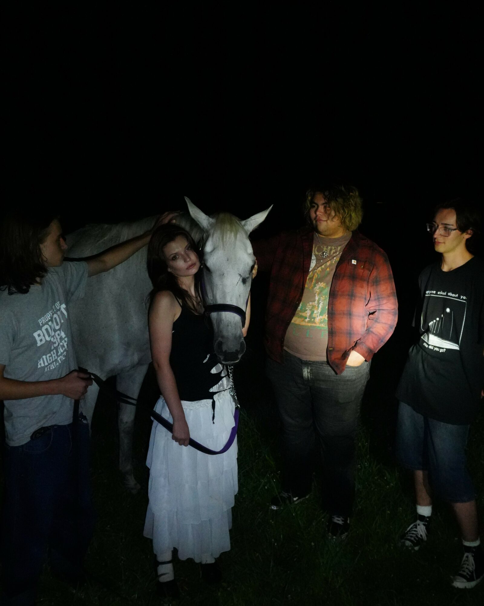 Four people and a white horse outdoors at night, with grass visible at the bottom.