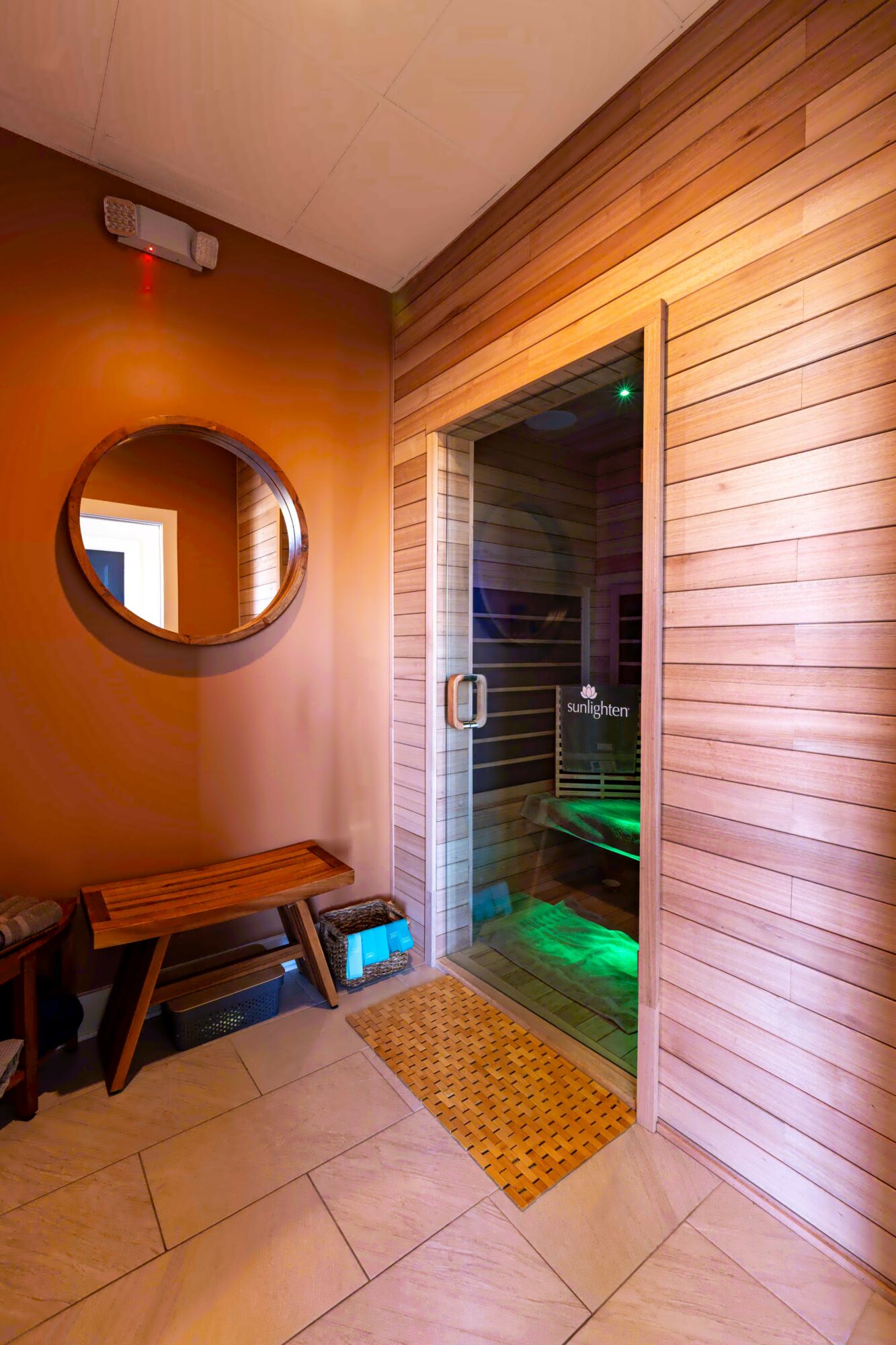 Sauna room with glass door, wooden walls, round mirror, bench, and a small mat outside the door.