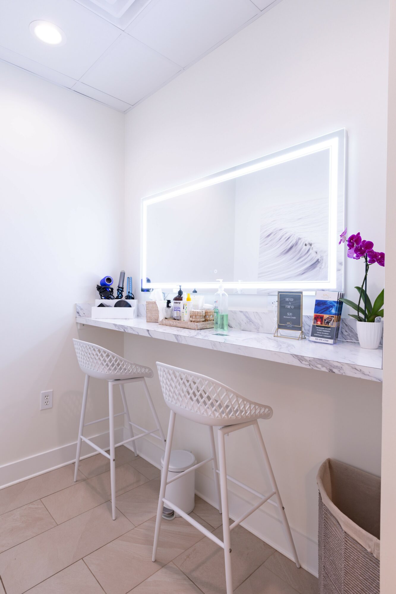 Bright vanity area with illuminated mirror, white countertop, two white chairs, and a potted purple orchid.