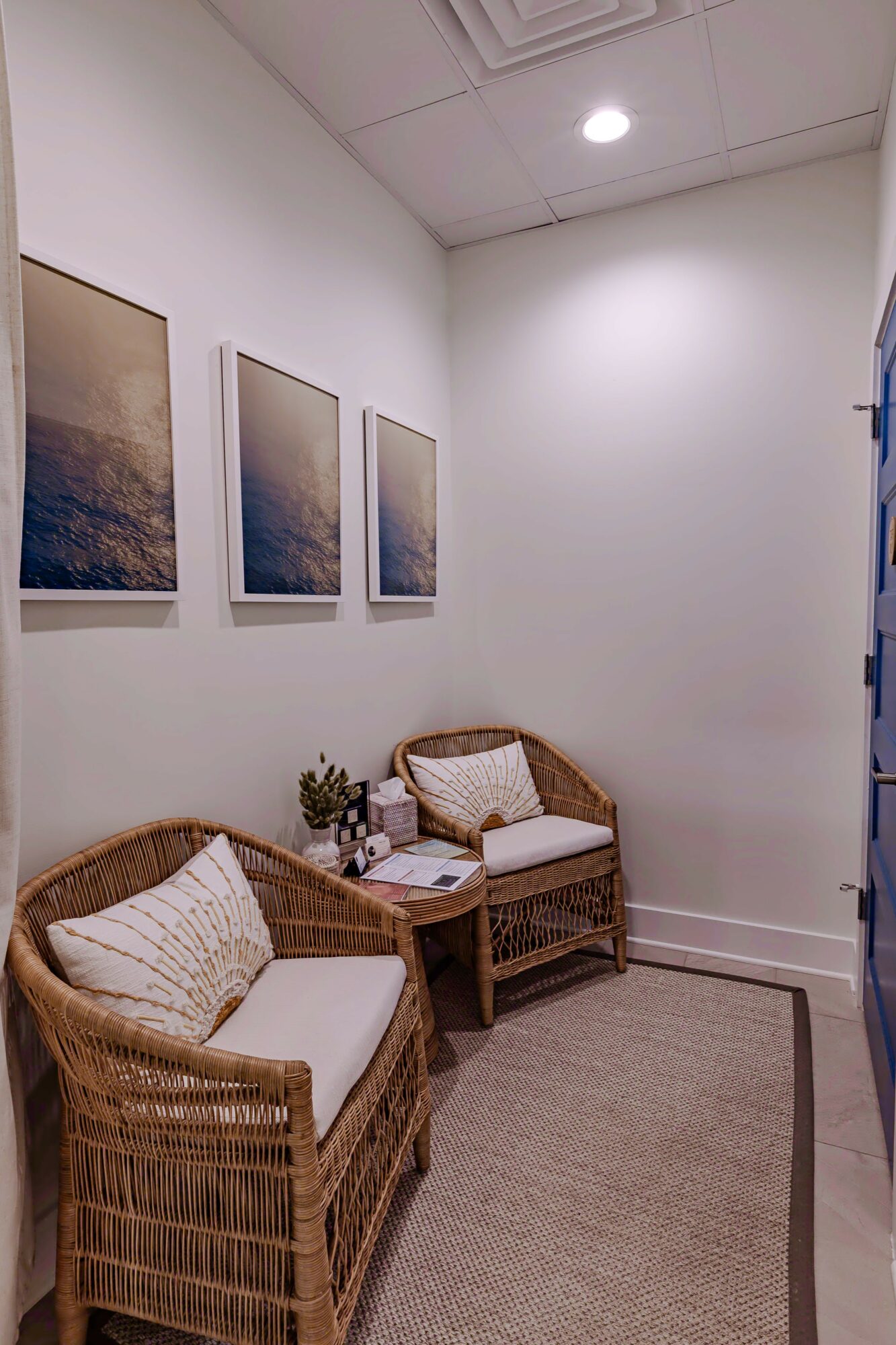 Corner of a room with two wicker chairs, a small table, framed artwork, and a beige rug.