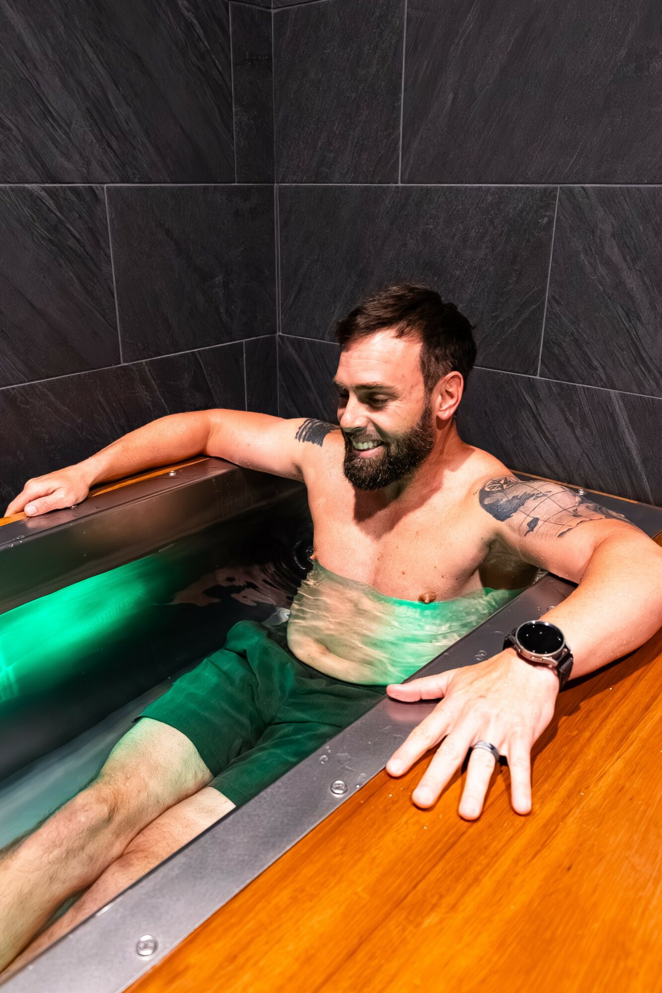 Man with a beard relaxing in a bathtub with dark tiled walls, smiling, wearing a watch, and resting his arm on the edge.