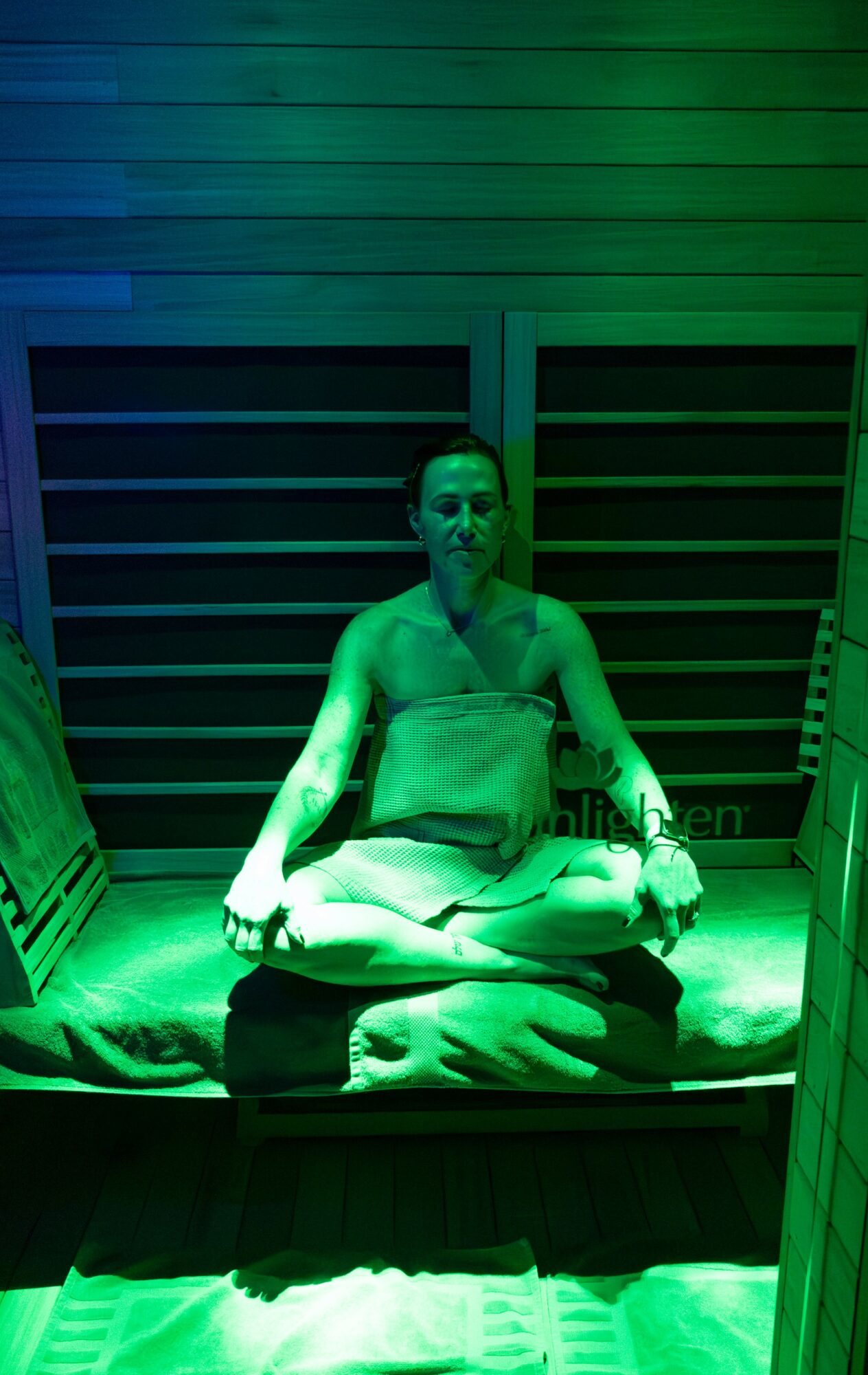 Person sitting cross-legged on a platform, illuminated by green light, with a wooden slatted wall behind, in a meditative pose.
