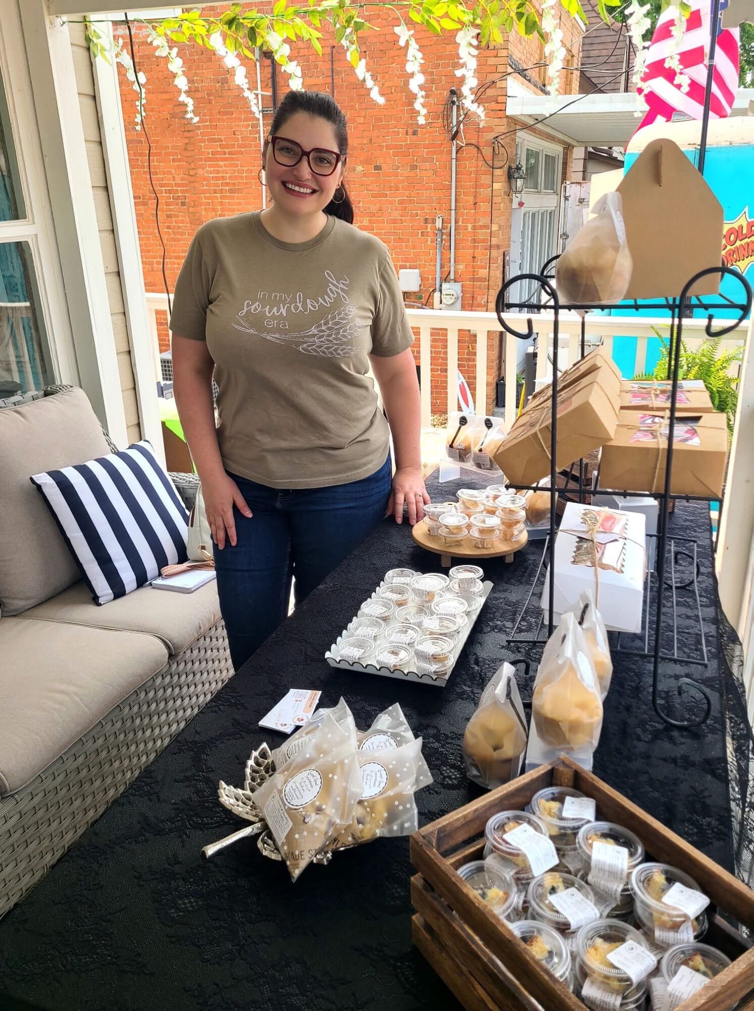 Woman standing behind a table with baked goods, smiling, on a porch with outdoor furniture and decorations.