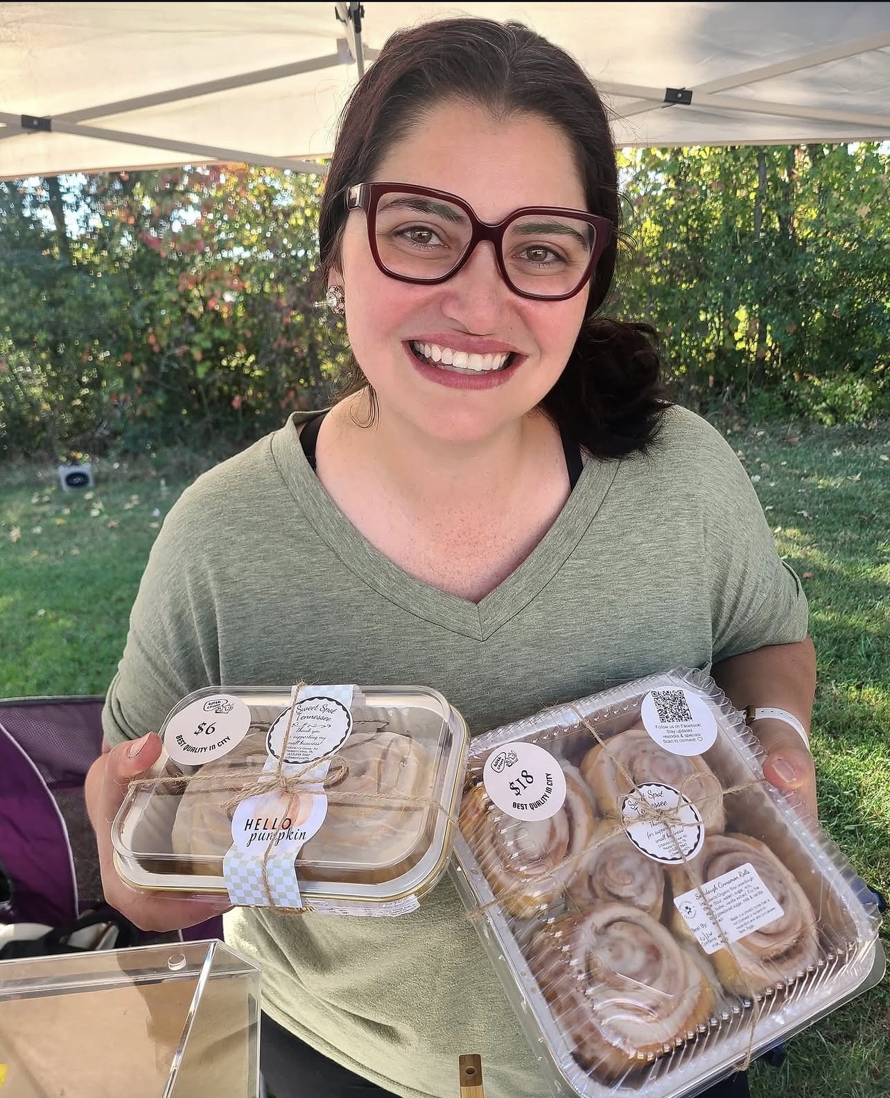 Smiling woman with glasses holding two trays of baked goods outdoors under a canopy, with trees and grass in the background.