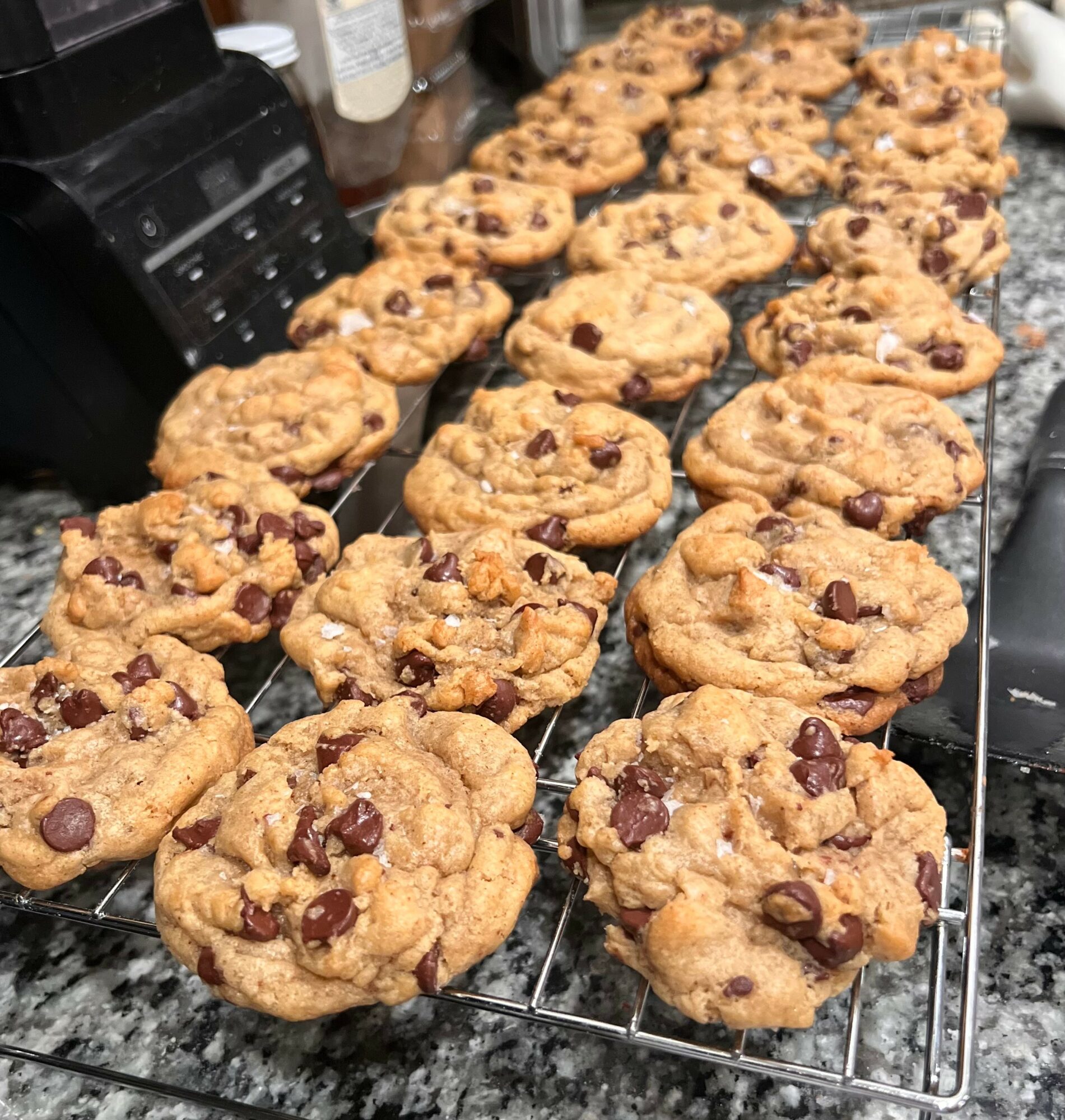 Chocolate chip cookies cooling on a wire rack, with a kitchen counter and appliances in the background.