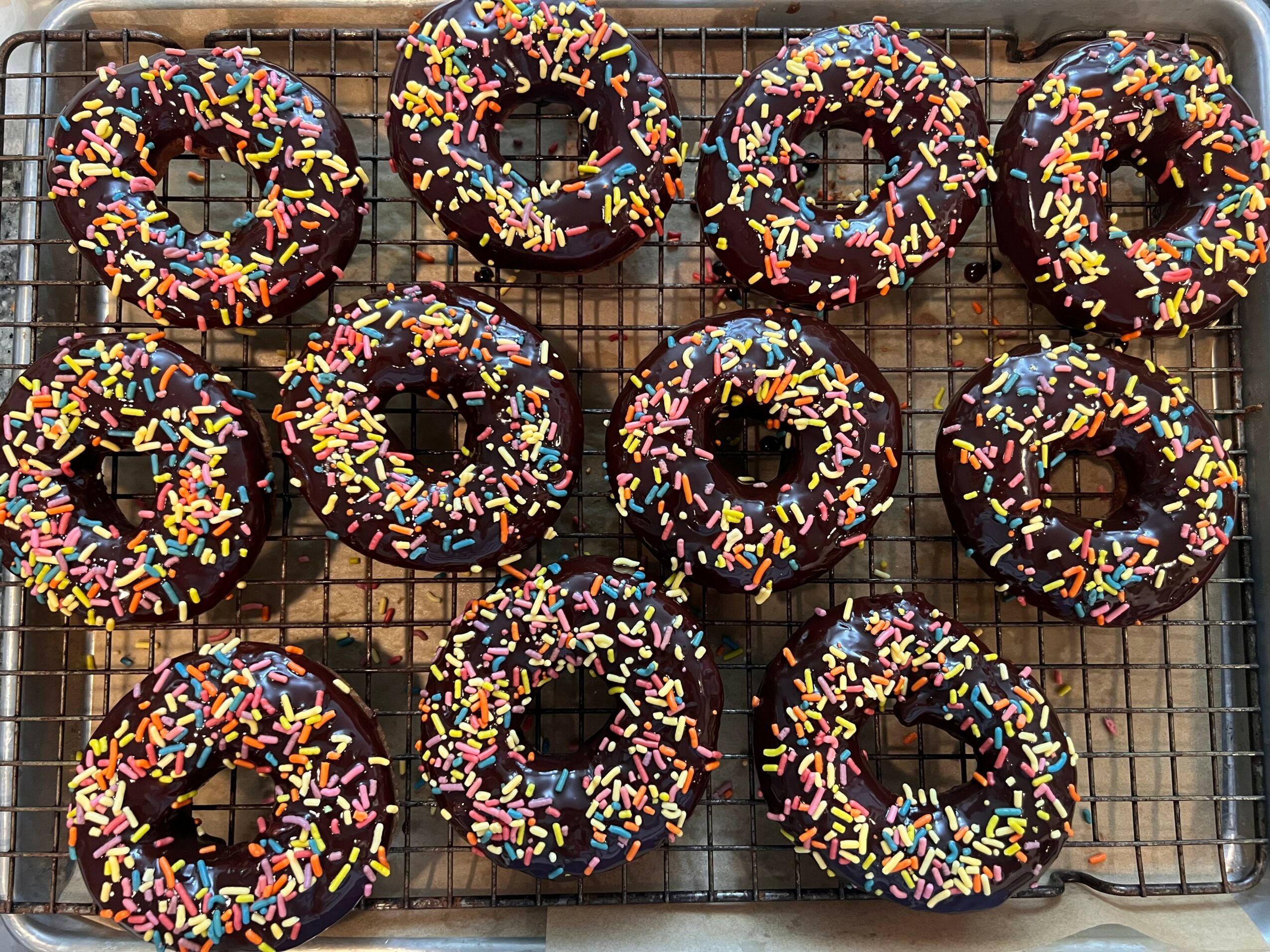 Ten chocolate donuts with colorful sprinkles on a wire cooling rack.