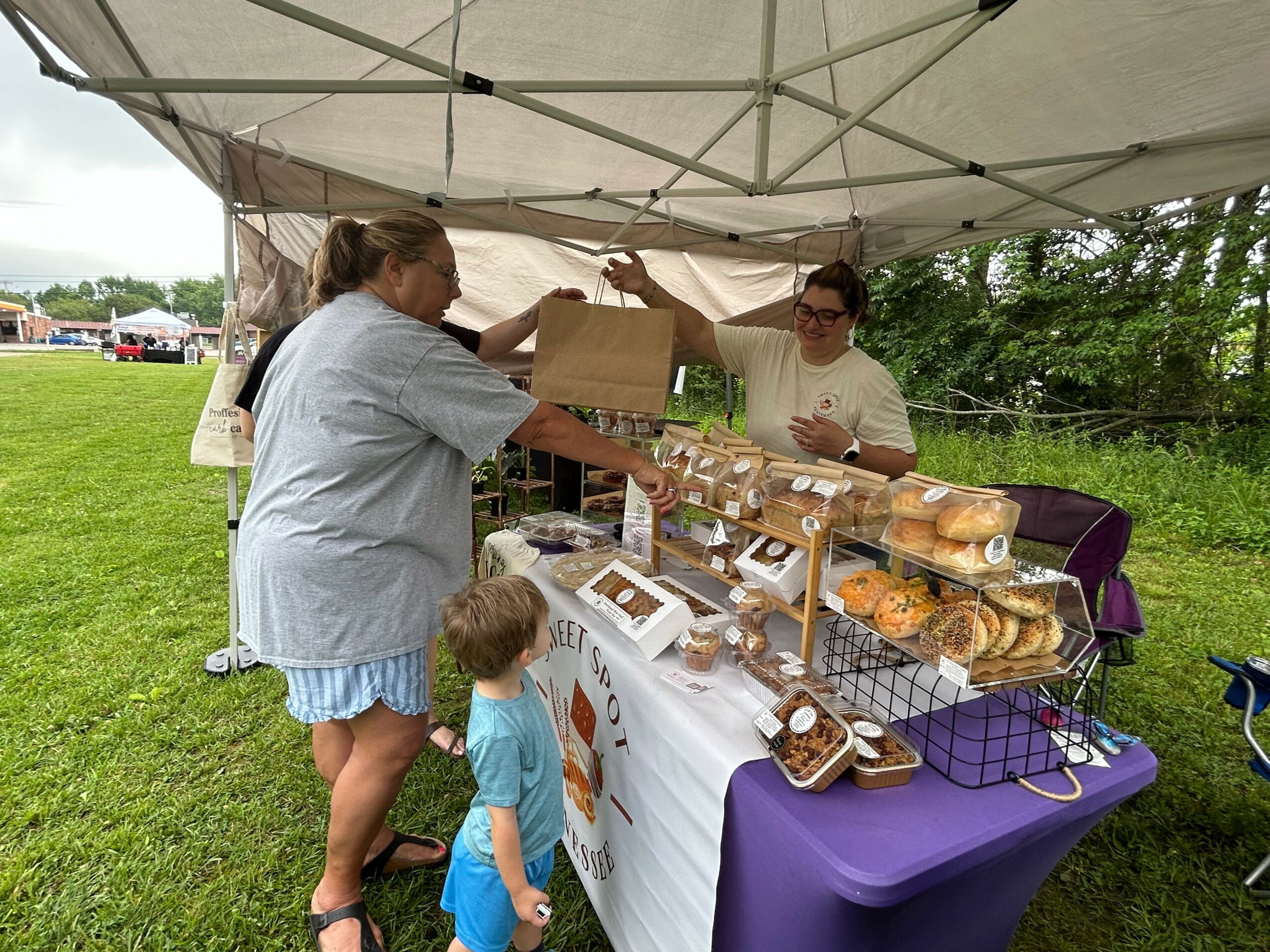 Two women and two children at an outdoor market stall with baked goods under a canopy, grassy area in background.