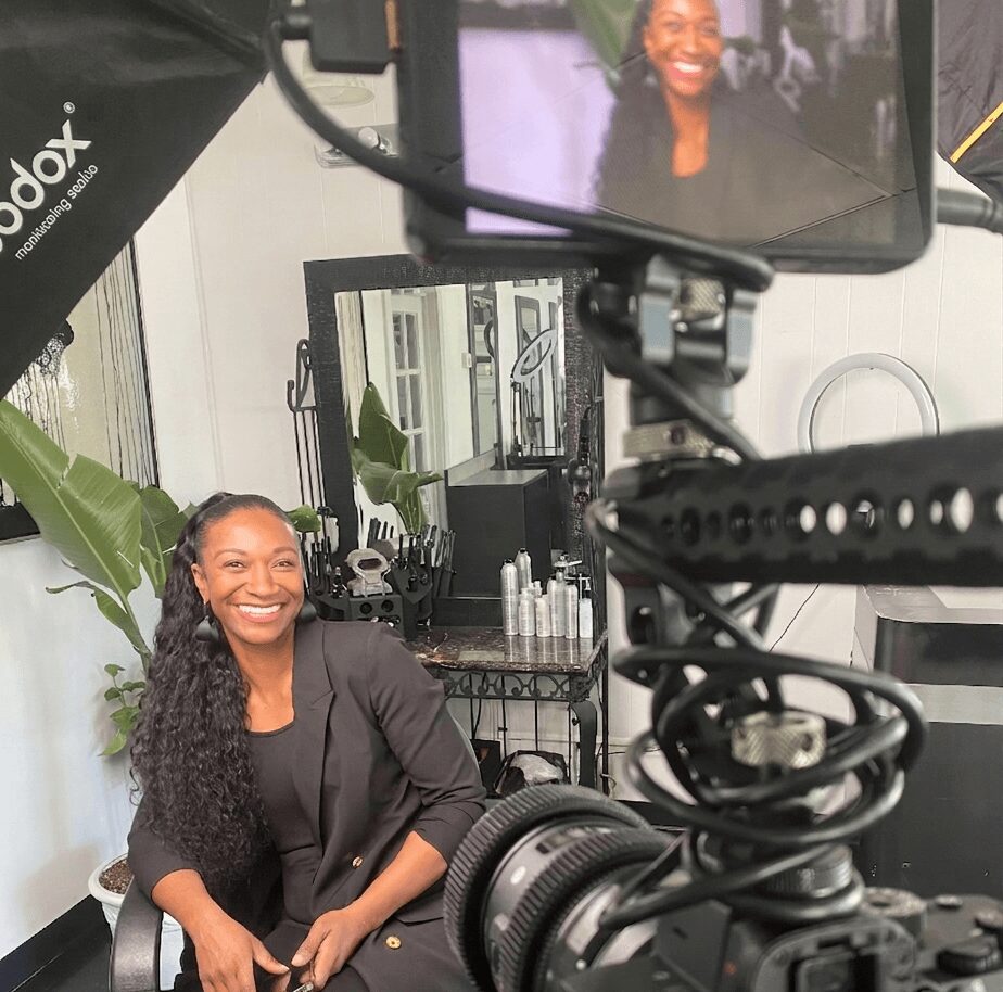 Woman with long curly hair smiling, sitting in a chair, in a room with plants and mirrors, camera setup visible.