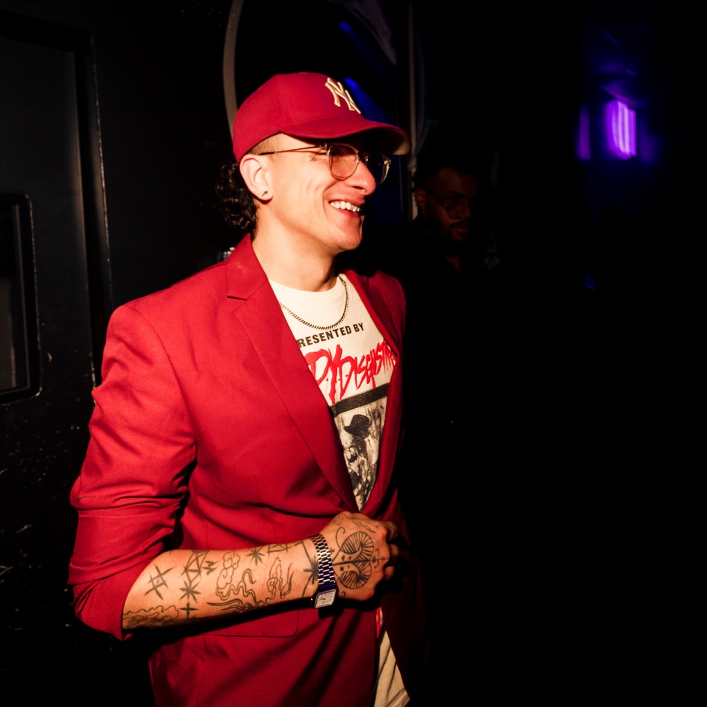 Person smiling, wearing a red blazer, baseball cap, glasses, and showing tattoos on arm, in a dark setting.