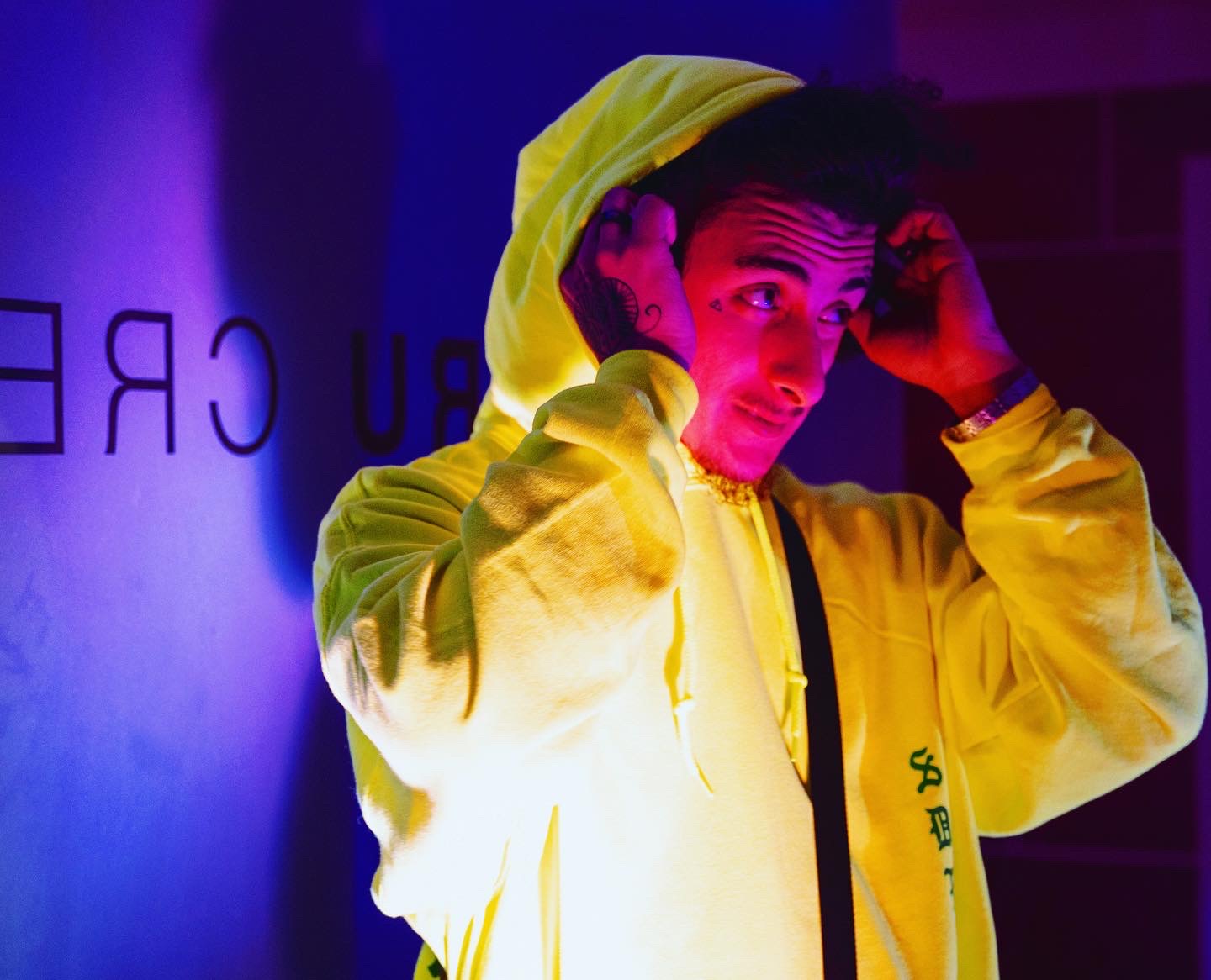 Young man in yellow hoodie adjusting hood, illuminated by colorful lighting, with partially visible text in background.