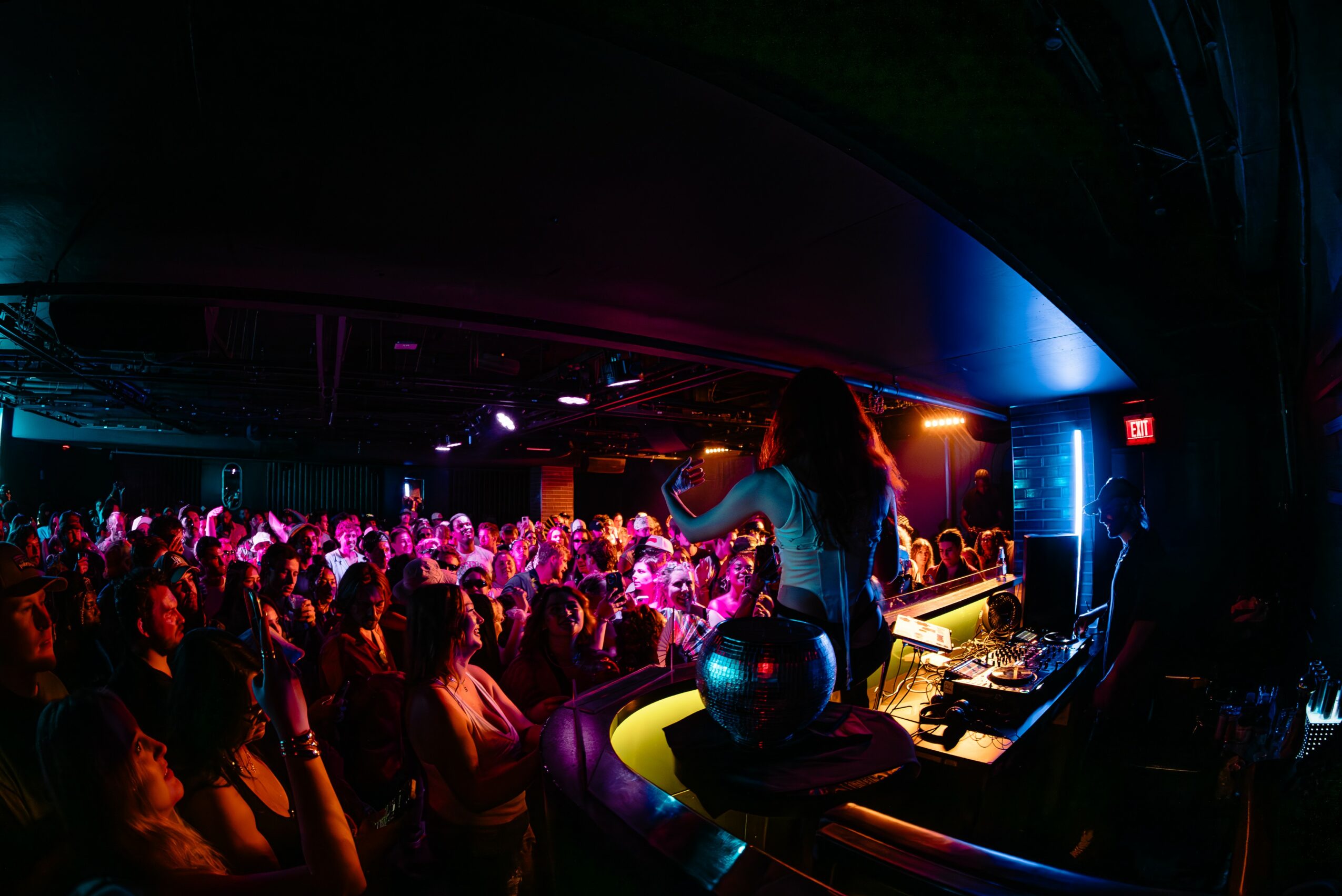Crowd of people in a dark club with colorful lighting, facing a performer on stage, with DJ equipment visible.