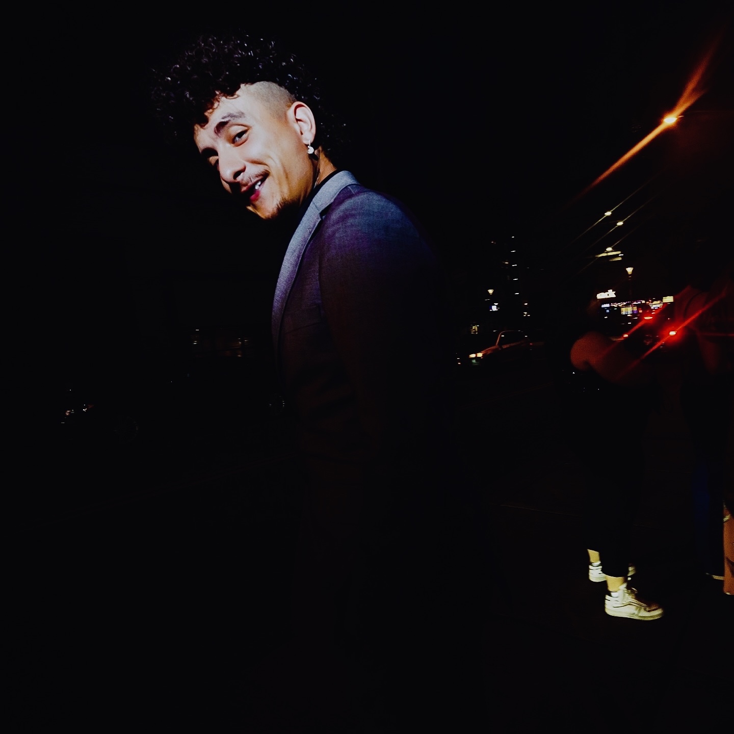 Young person with curly hair smiling, wearing a dark blazer, in a dark environment with bright lights in the background.