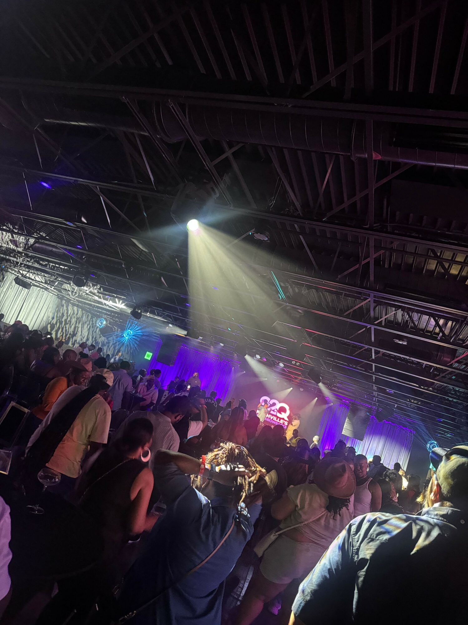 Crowd of people at a concert or event with stage lighting and beams overhead.