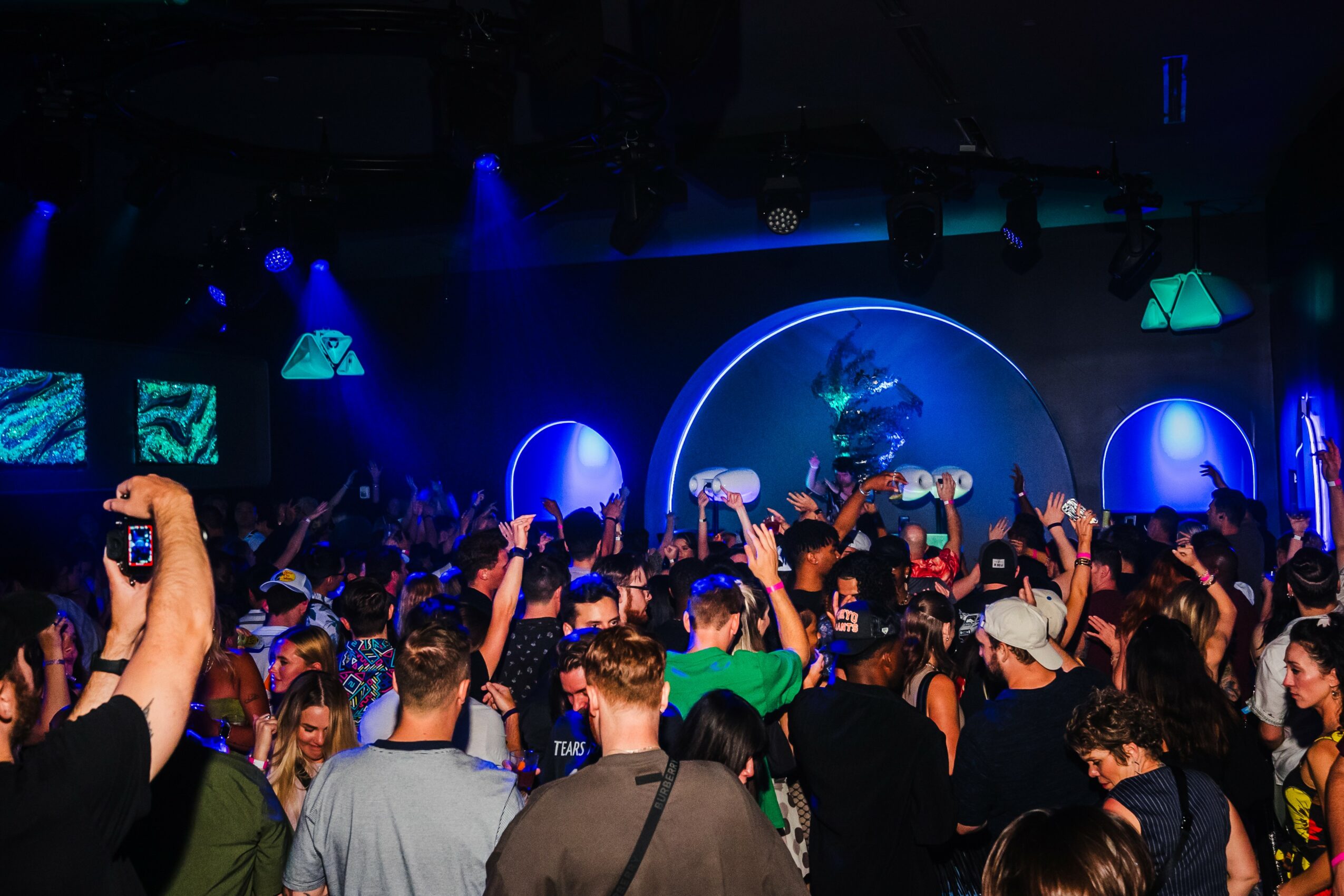 Crowd of people at a concert or event with a stage, blue lighting, and circular screens, some raising hands.