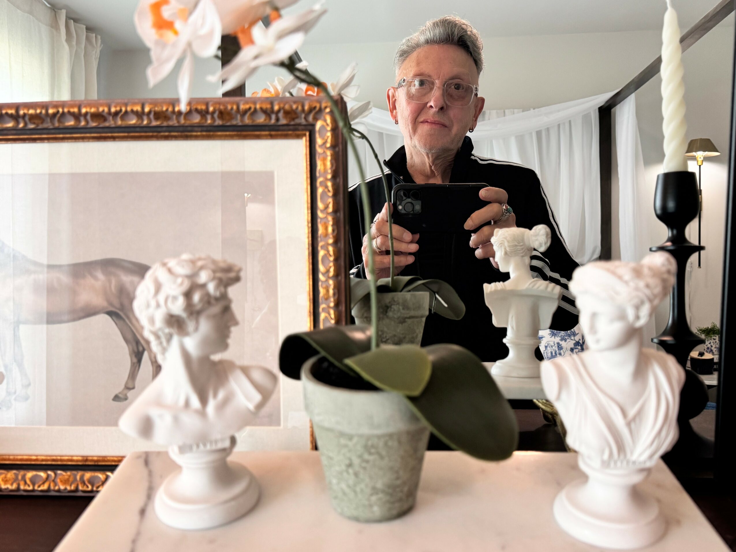 Man taking a selfie in a mirror, surrounded by classical busts, a framed artwork, and candles on a table.