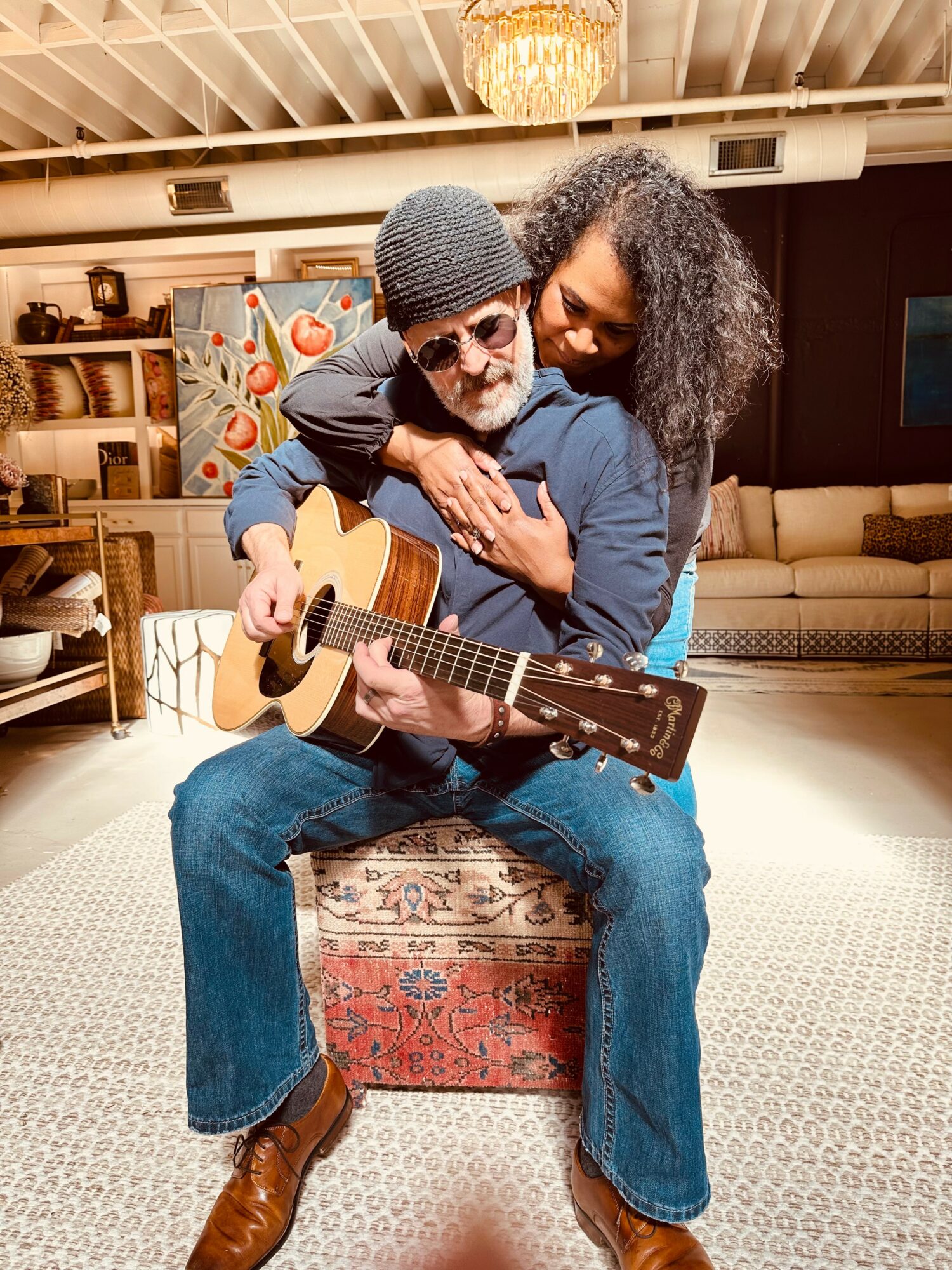 A man with a guitar sits on a chest, with a woman hugging him from behind in a cozy room.