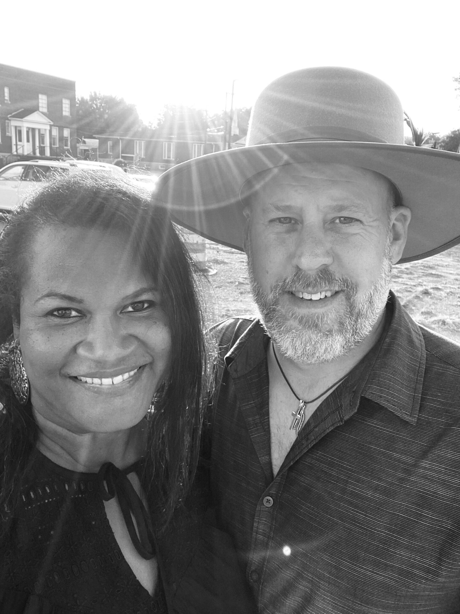 Two people smiling outdoors, one wearing a wide-brimmed hat, sunlight behind them, in black and white.