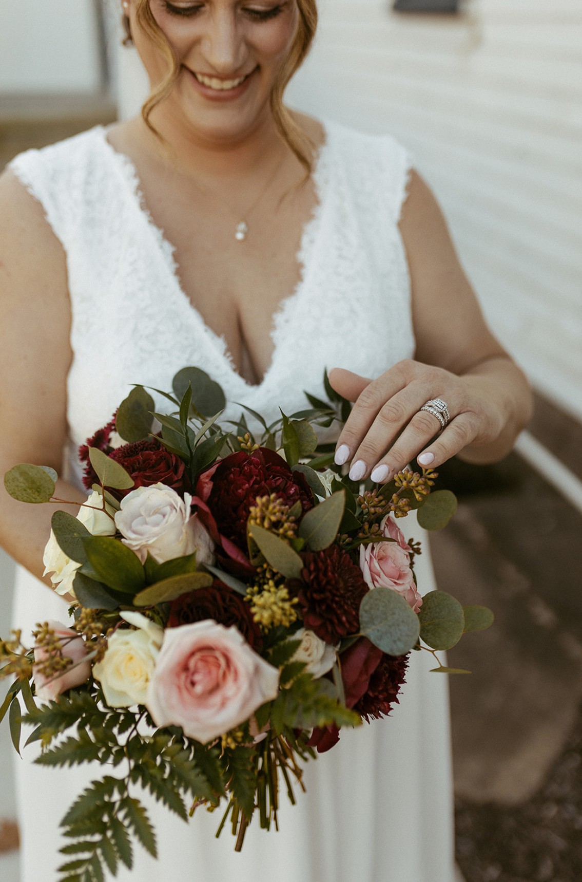 Woman in wedding dress holding a bouquet of roses, greenery, and flowers, smiling, outdoors.
