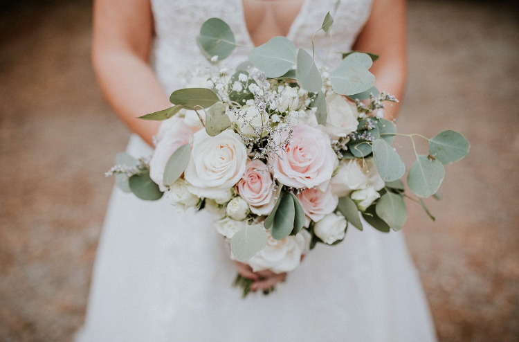 Person in a white dress holding a bouquet of white and pink roses with green leaves, outdoors.